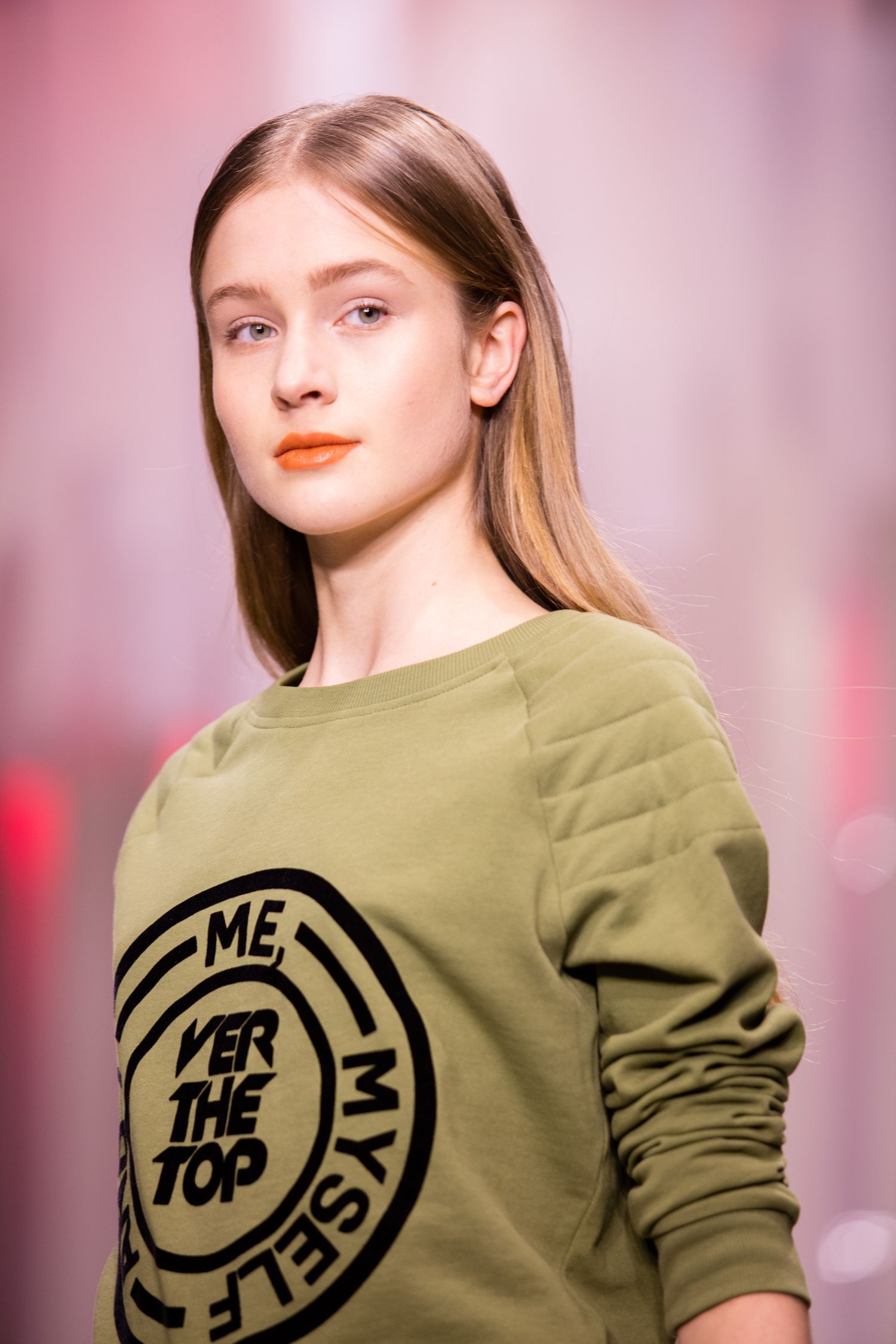 Young woman in olive green graphic sweatshirt reading 'Me, Myself, Over The Top' posing against pink background.