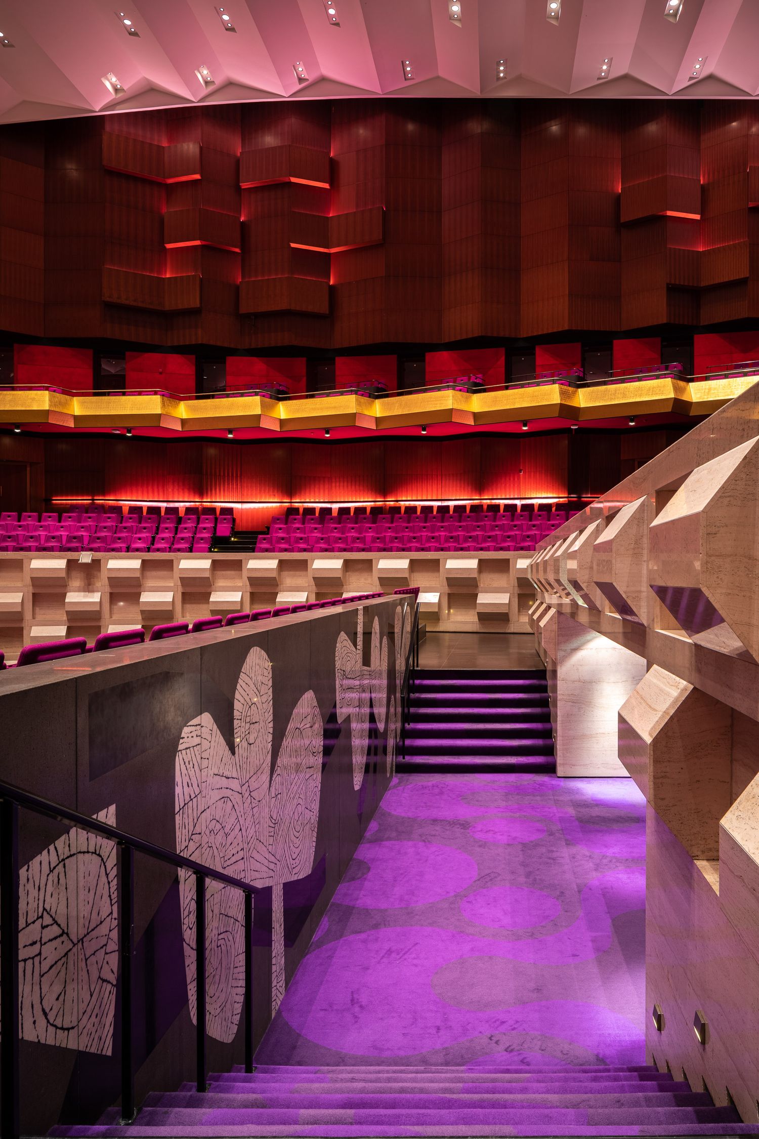 Modern theater interior with purple-lit staircase, red acoustic panels, and tiered seating in vibrant colors.