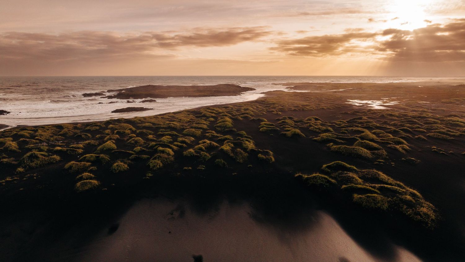 Aerial view of a dramatic coastal landscape with mossy rocks and golden sunset over the ocean horizon.