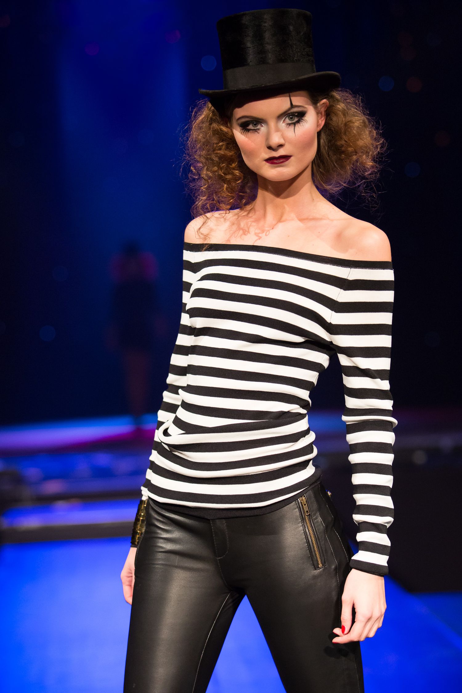 Model on runway wearing black and white striped off-shoulder top, leather pants, and black top hat with dramatic makeup.