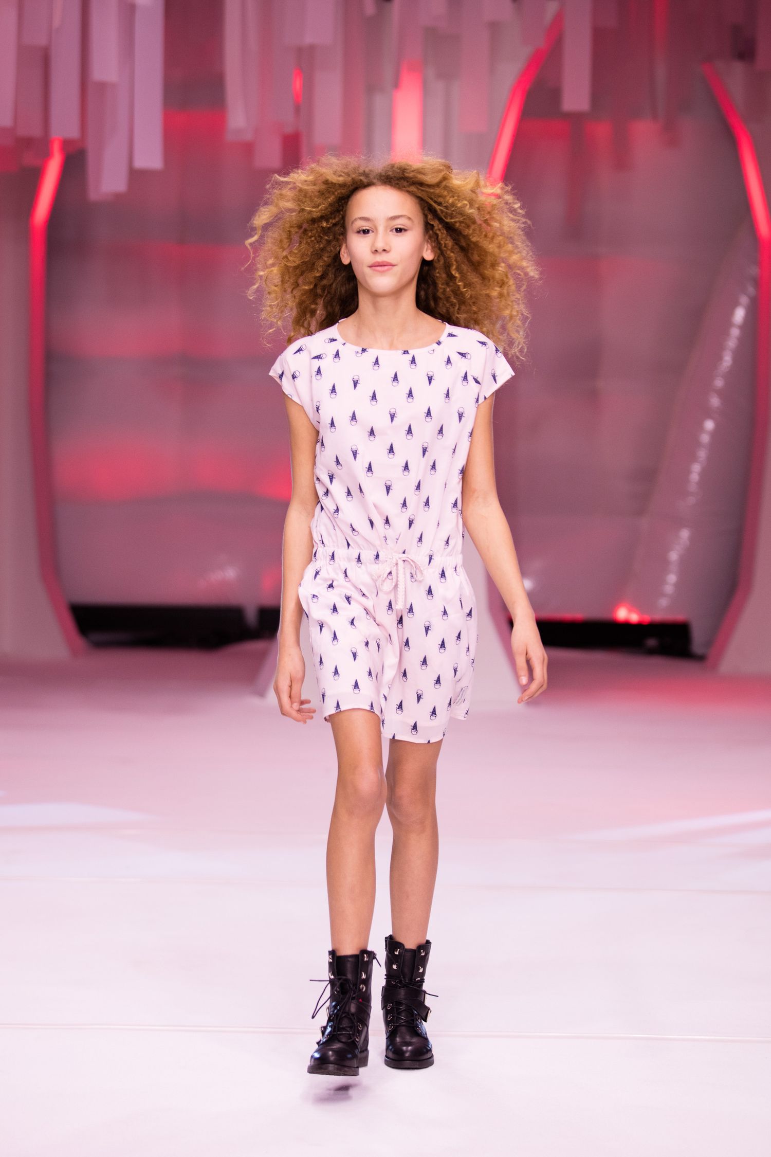 Young girl model walks runway in white printed romper with black boots at a children's fashion show.