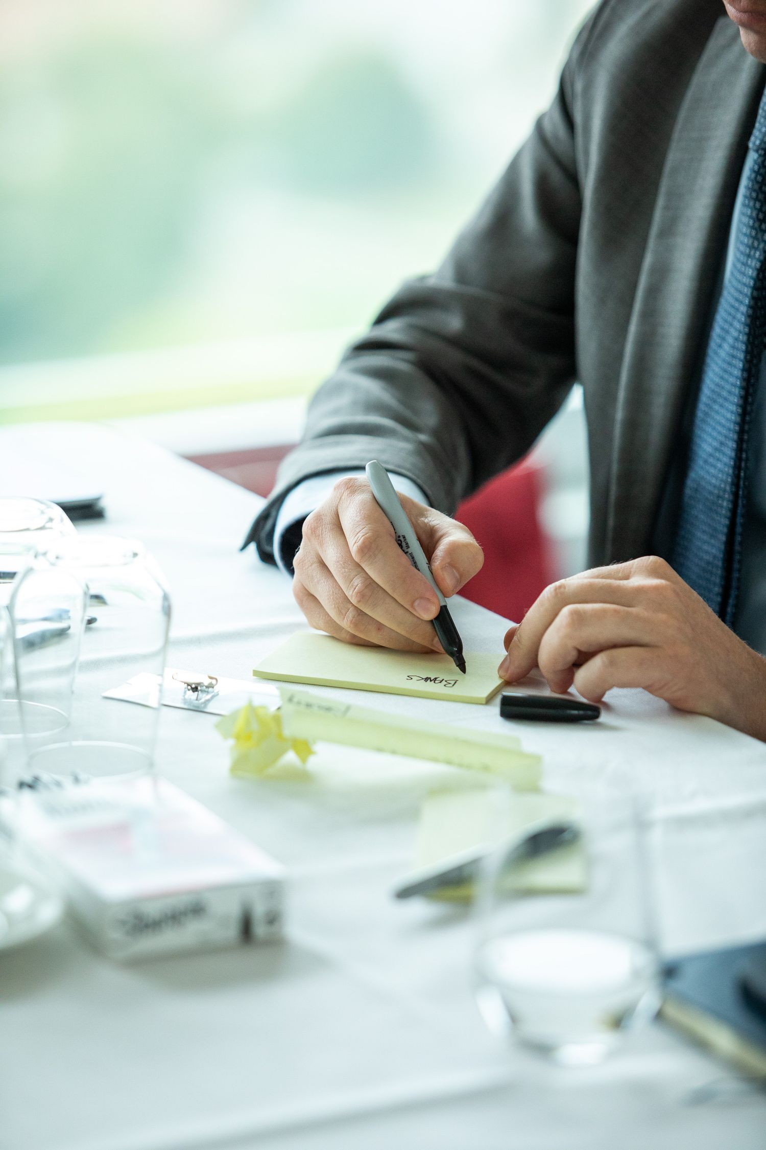 Professional in business attire writes notes at desk with glass of water and office supplies in bright setting.