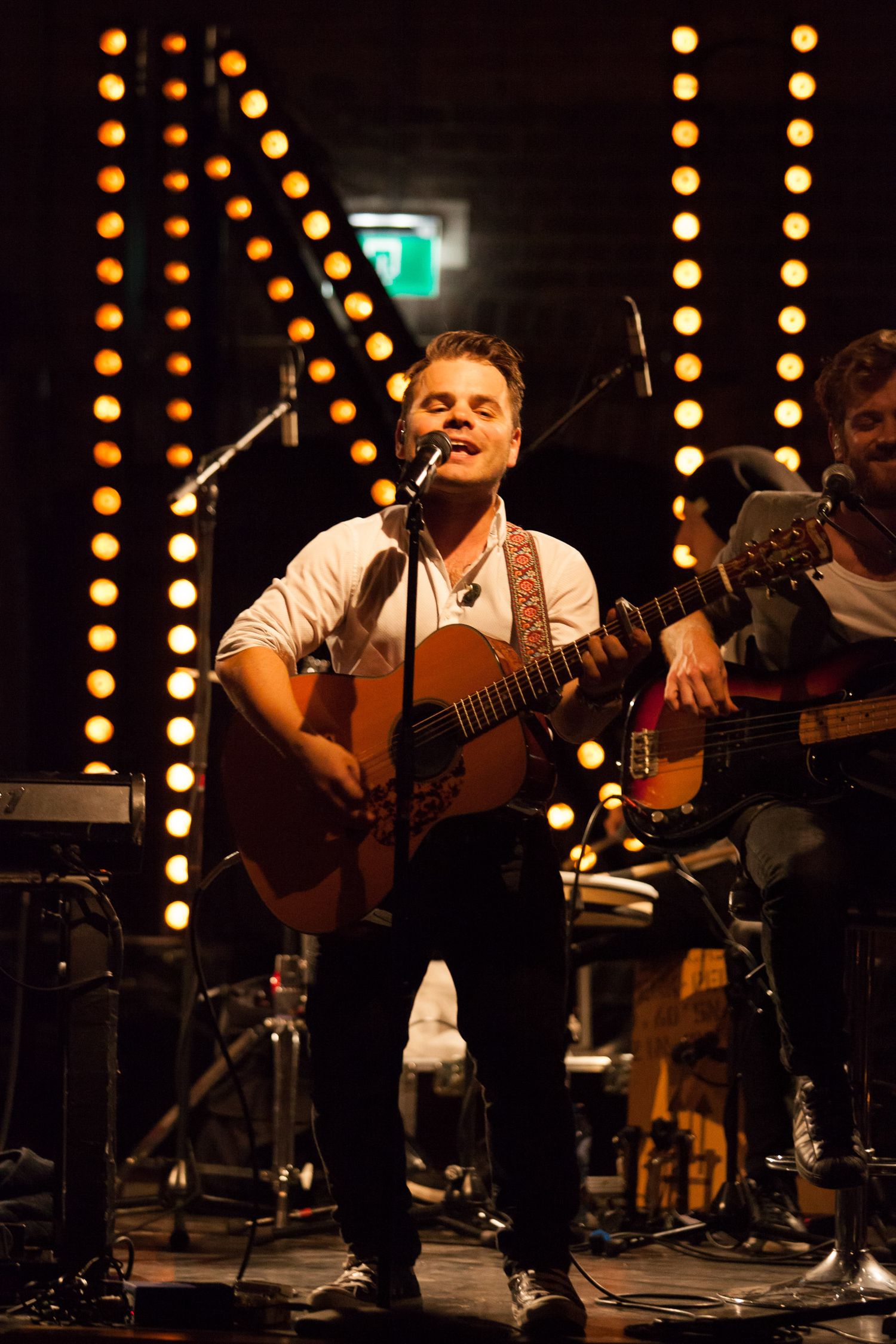 Male musician plays acoustic guitar and sings into microphone on stage with warm yellow lights behind him.