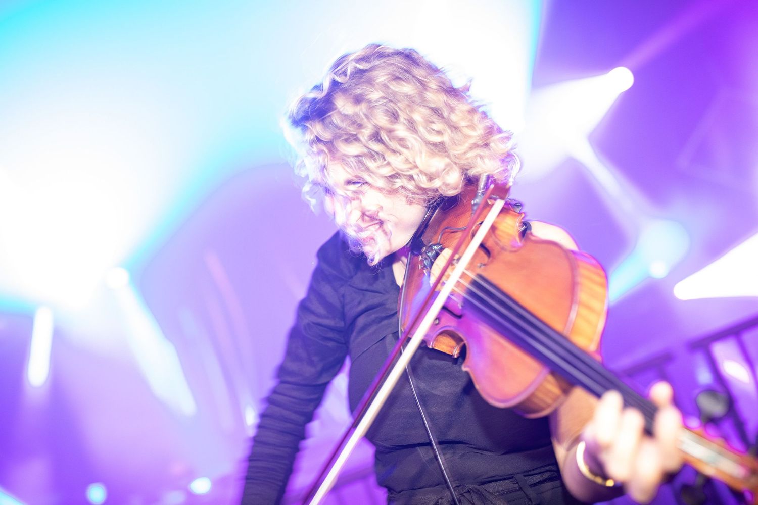 A guitarist performs on stage with colorful purple and blue lighting effects during a live concert performance.