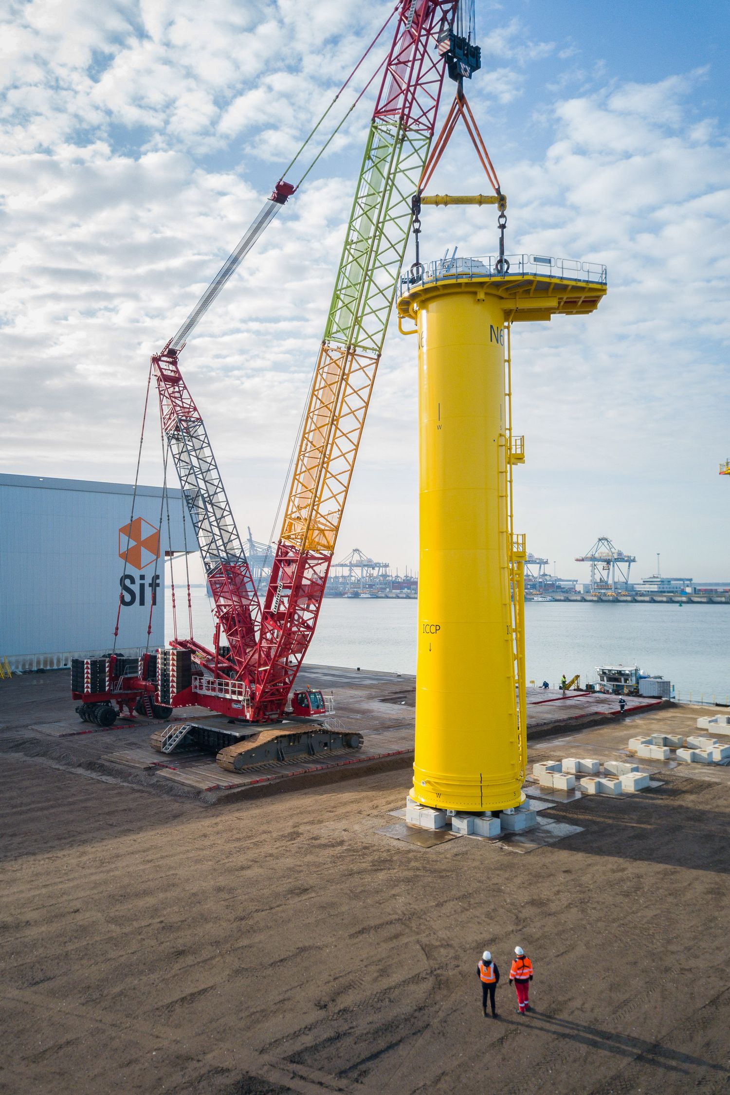 Yellow wind turbine tower sections being installed with multiple cranes at a coastal site.