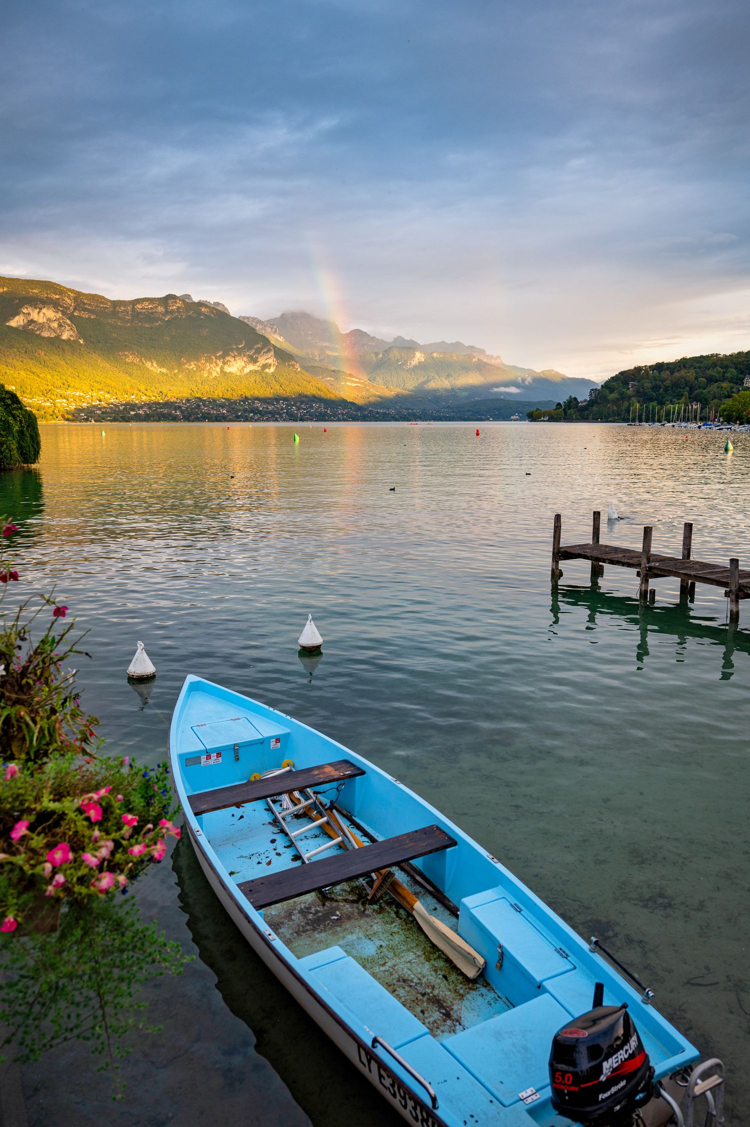 Blue wooden boat moored at wooden pier on Lake Annecy with mountain views at sunset.