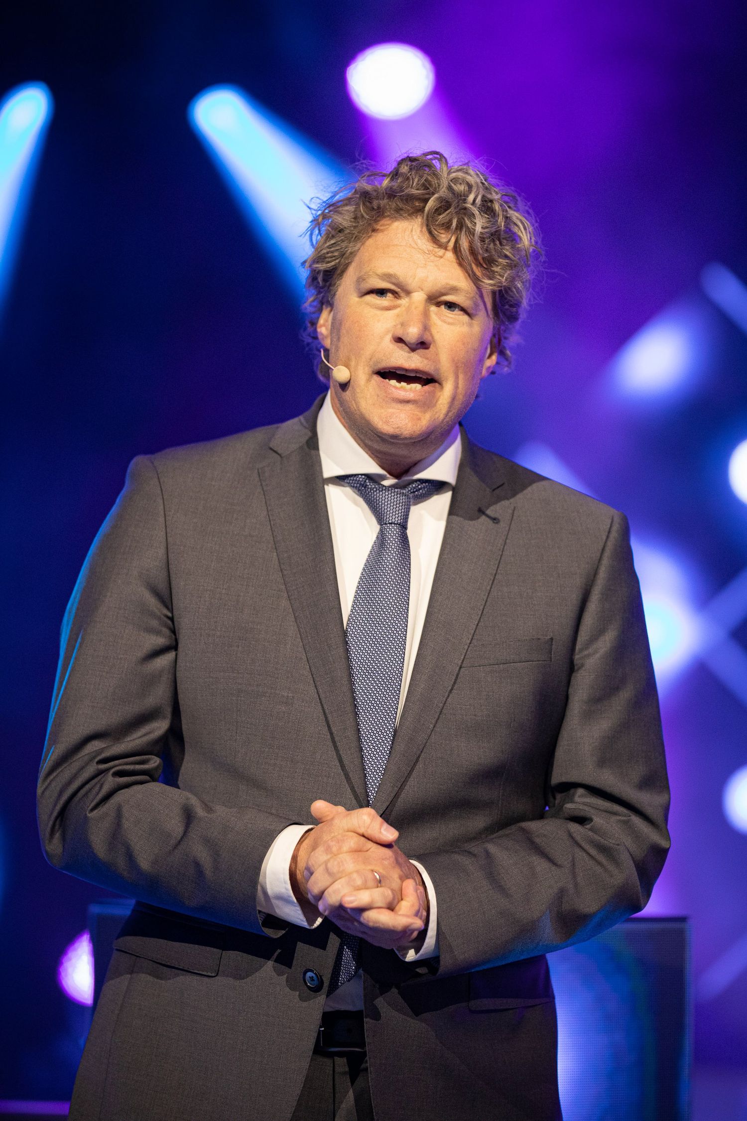 A performer in a grey suit gestures expressively on stage with dramatic purple and blue lighting in the background.