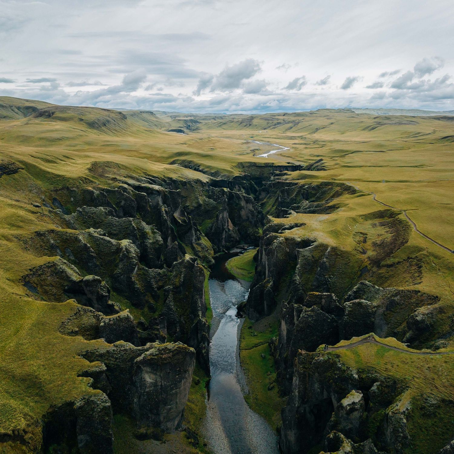 Aerial view of a deep river canyon winding through green rolling hills under a cloudy sky in Iceland.