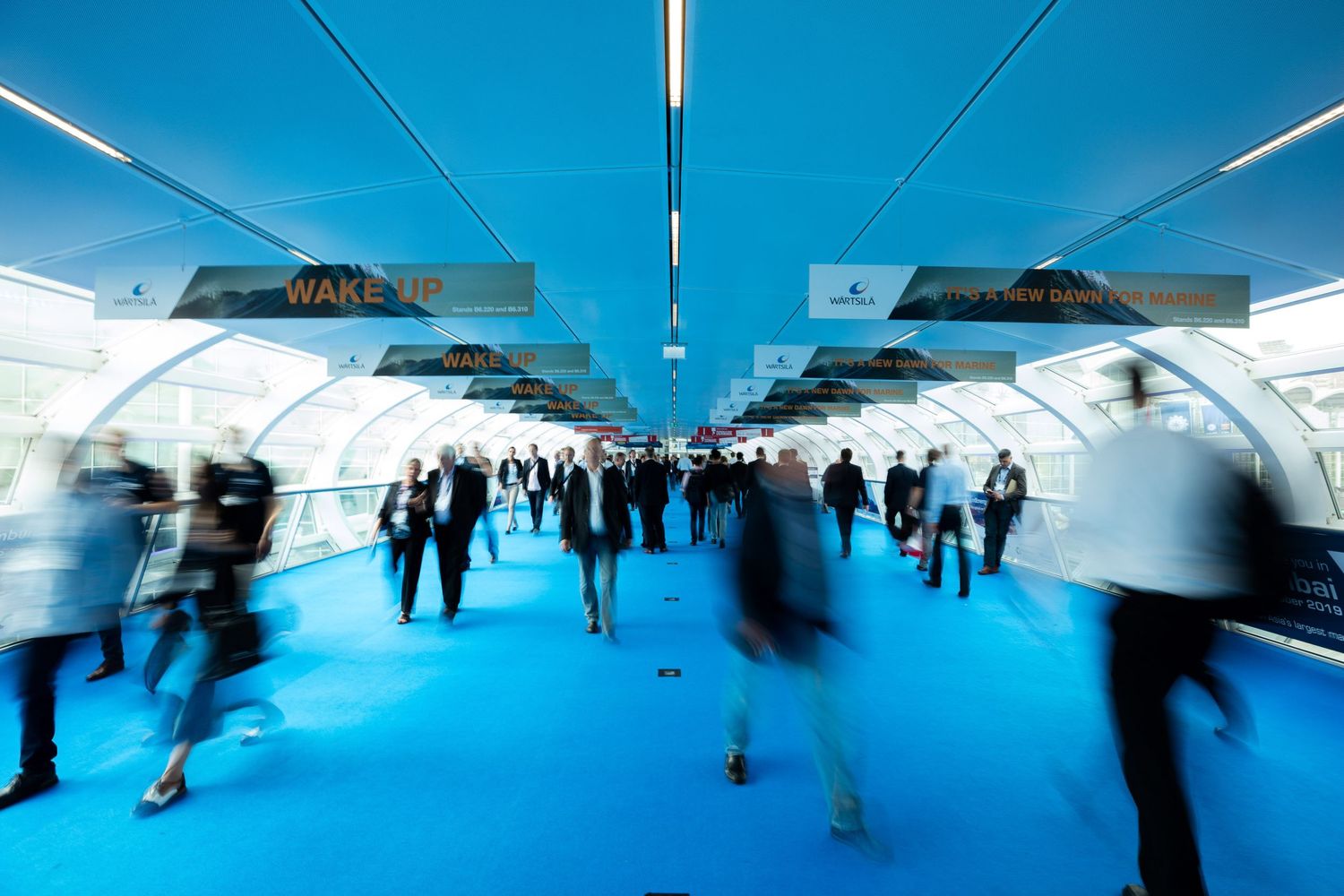 Commuters walk through a modern subway station corridor with bright blue lighting and reflective surfaces.