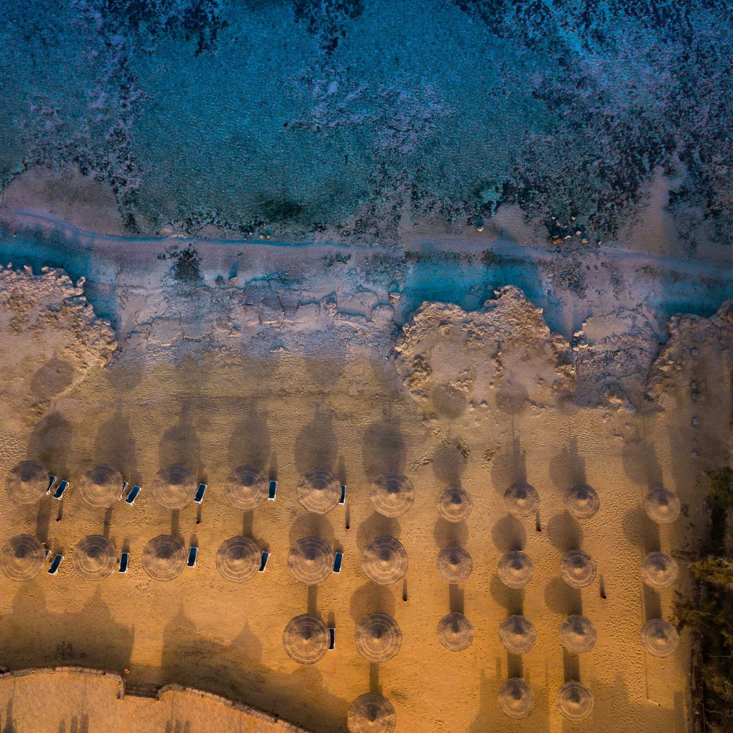 Aerial view of a sandy beach with turquoise ocean waves and rows of beach umbrellas casting shadows.