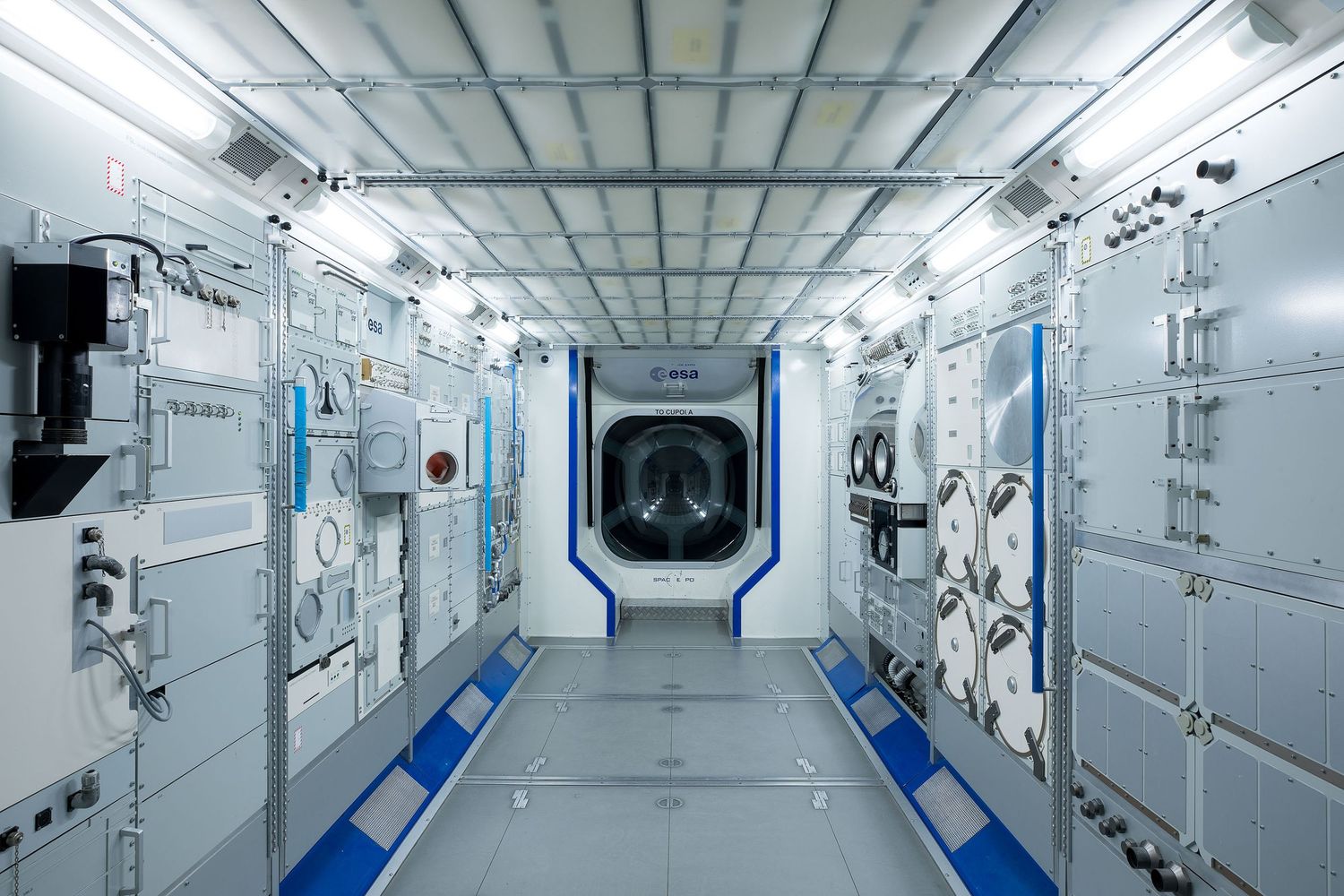 Interior corridor of a space station module with white panels, blue accents, equipment racks, spacesuits, and a hatch door.
