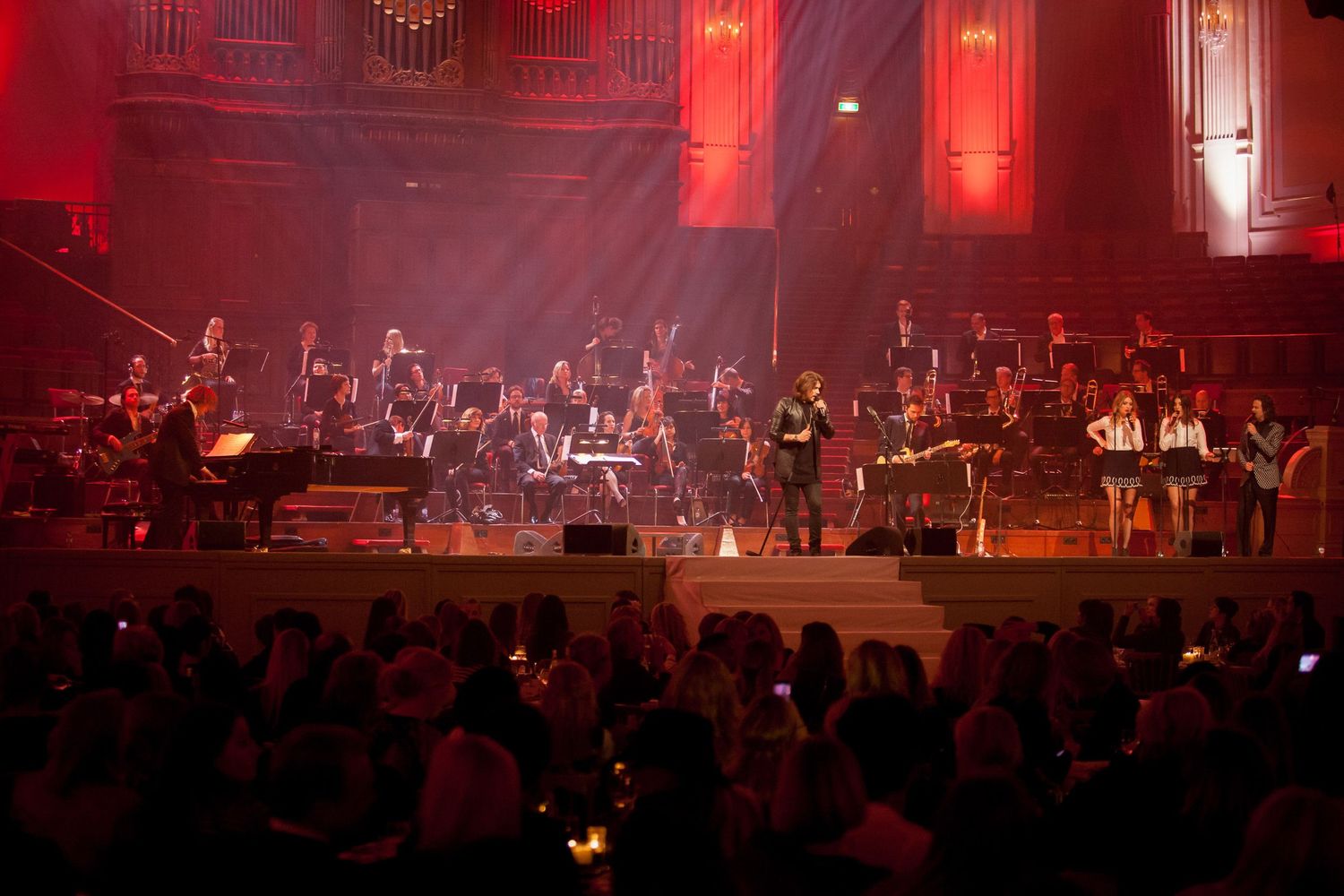 Orchestra performing on a grand stage with red lighting and a large audience in an elegant concert hall.