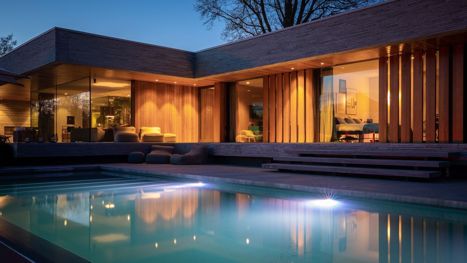 Modern luxury home with illuminated swimming pool and glass walls glowing warmly at twilight.