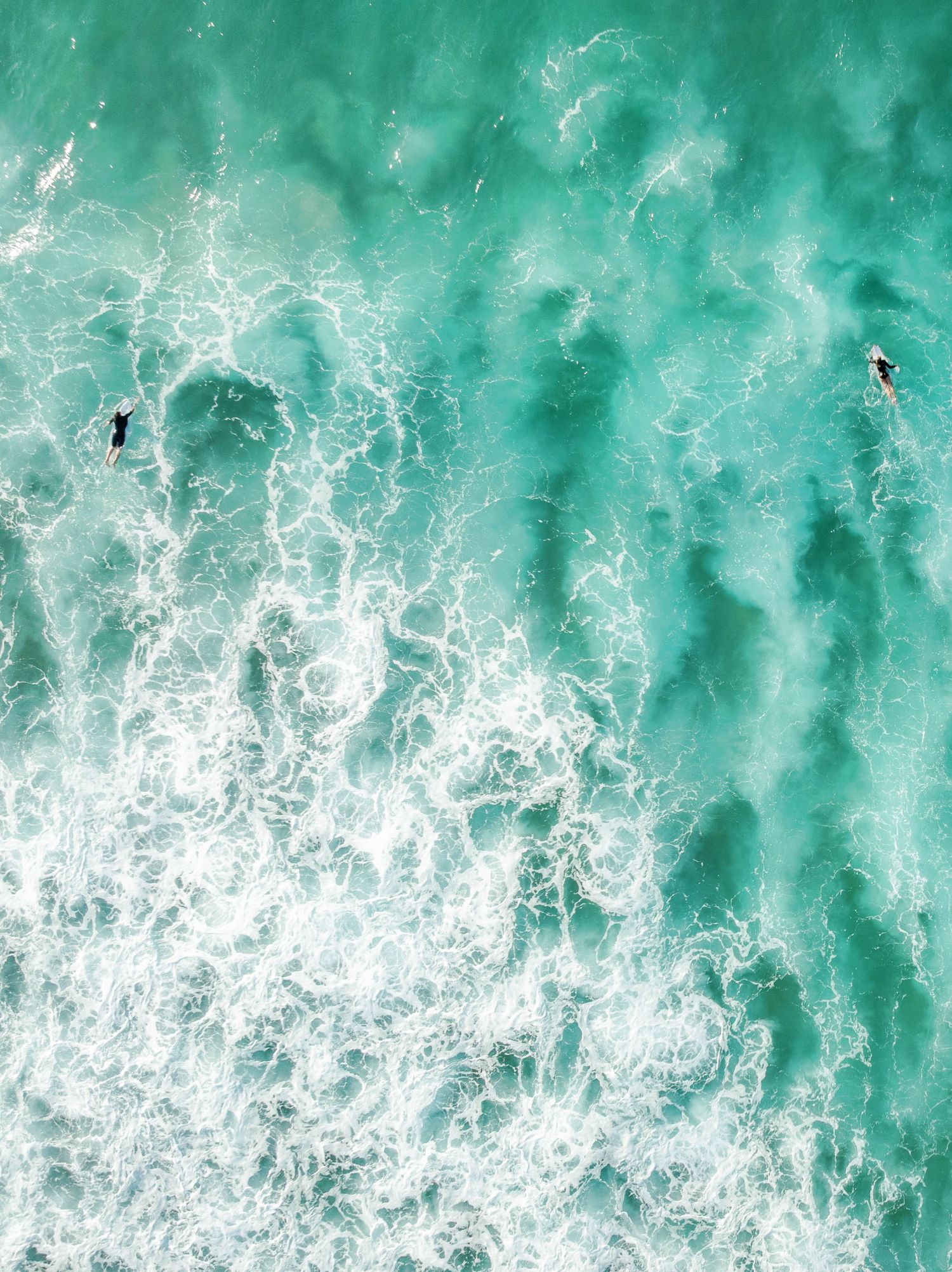 Aerial view of turquoise ocean waves with white sea foam and two surfers visible in the water.