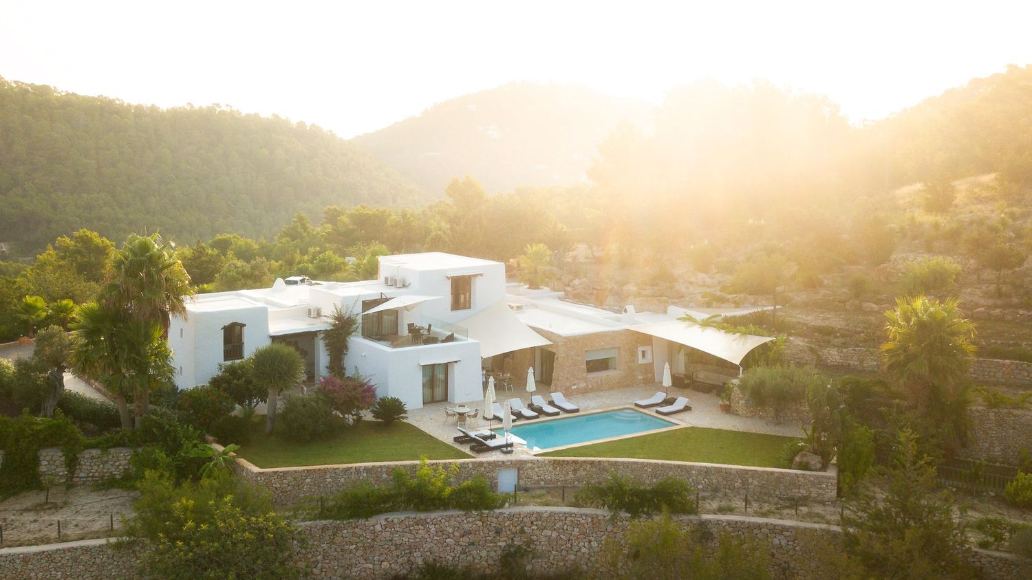 Aerial view of a modern white villa with a swimming pool surrounded by lush green hills bathed in golden sunlight.