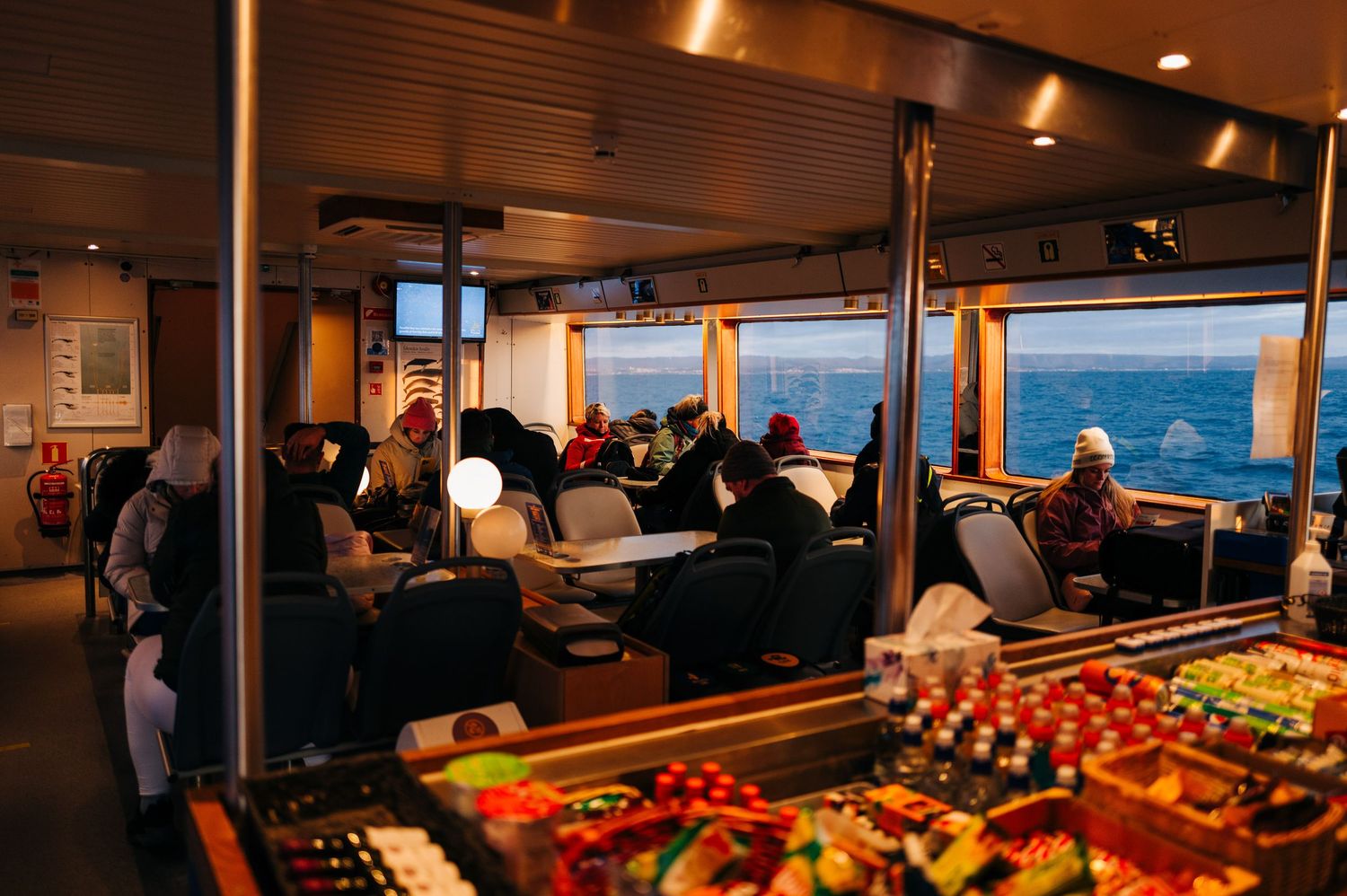 Passengers relax in comfortable seating aboard a cruise ship while colorful fruit displays are visible in the dining area.