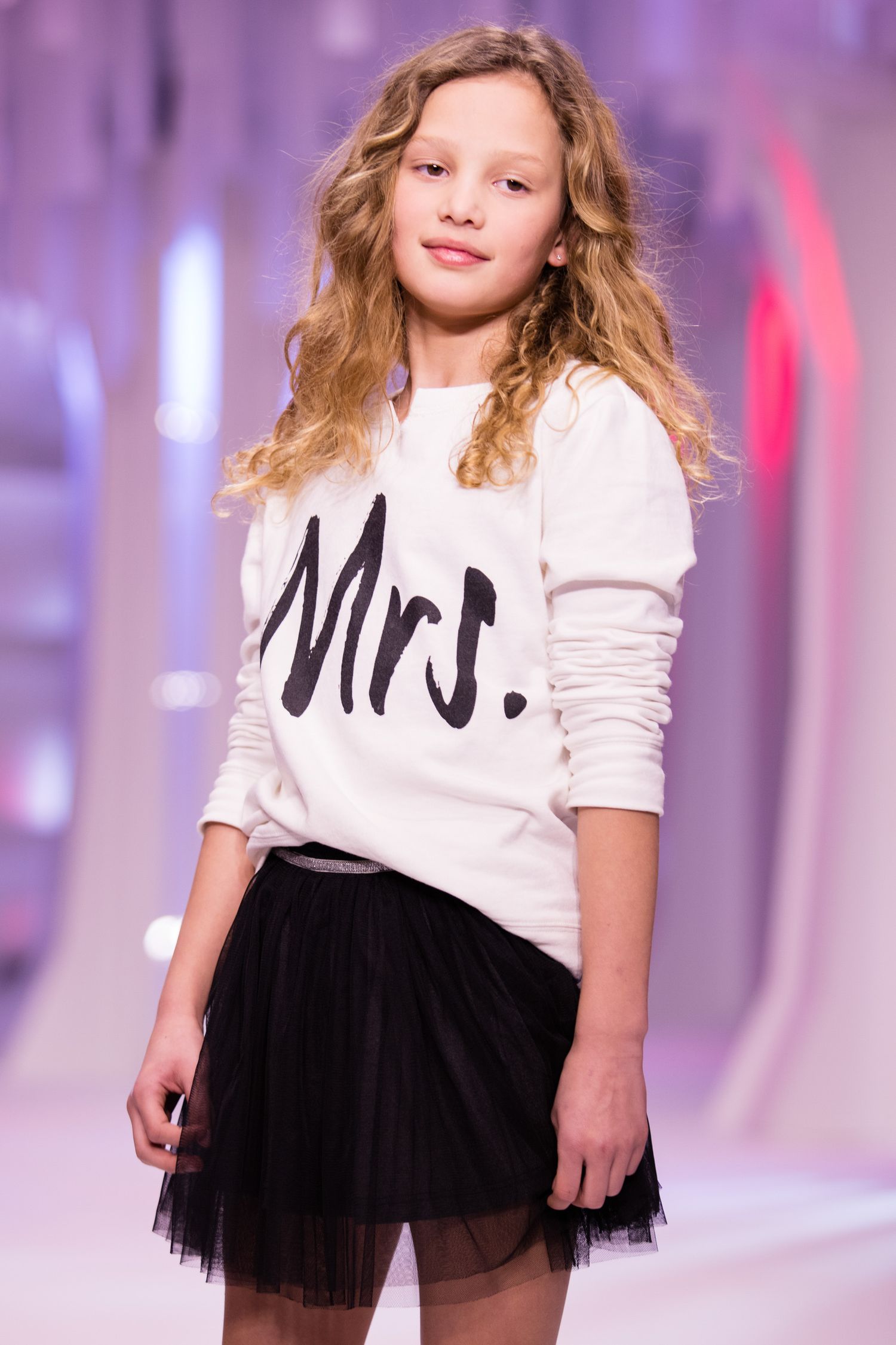 Young girl model on runway wearing white Mrs. sweatshirt and black tulle skirt at fashion show.