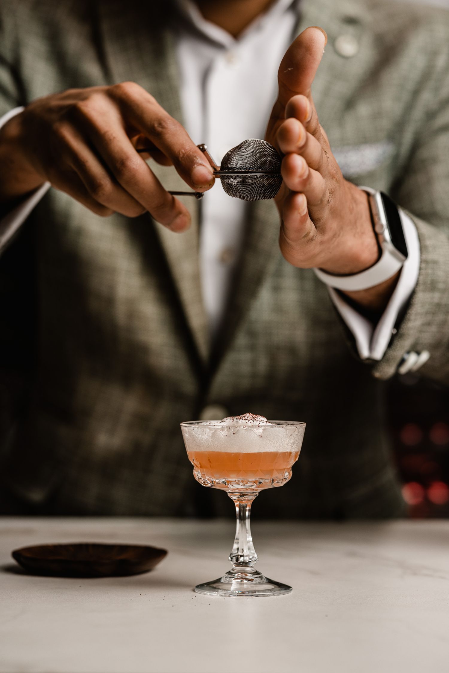 A bartender in a suit garnishes a pink cocktail in a coupe glass with a dark sphere on a white marble bar counter.