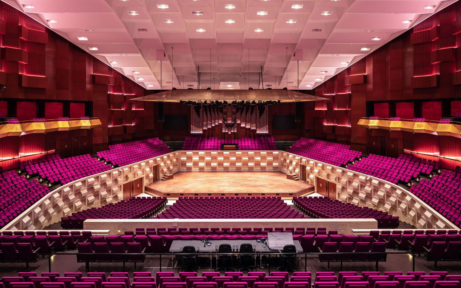 Modern concert hall interior with curved seating rows illuminated by pink lighting and featuring a wooden performance stage.