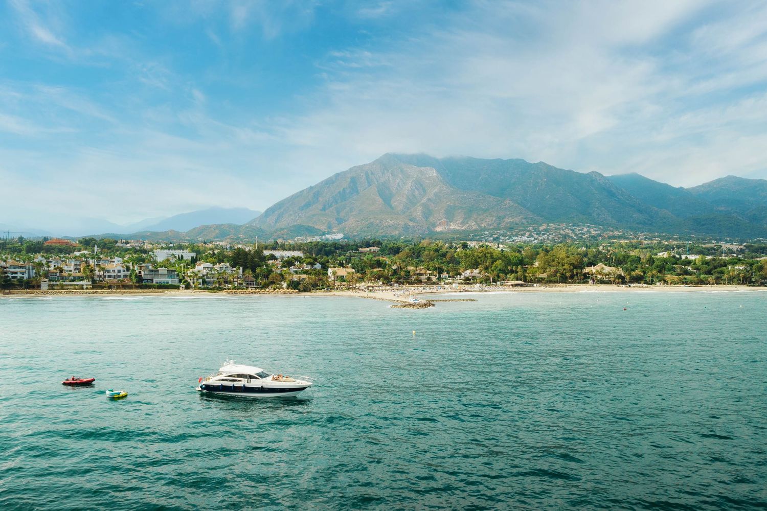 Aerial view of a coastal town with turquoise waters, boats, and mountains under a blue sky.
