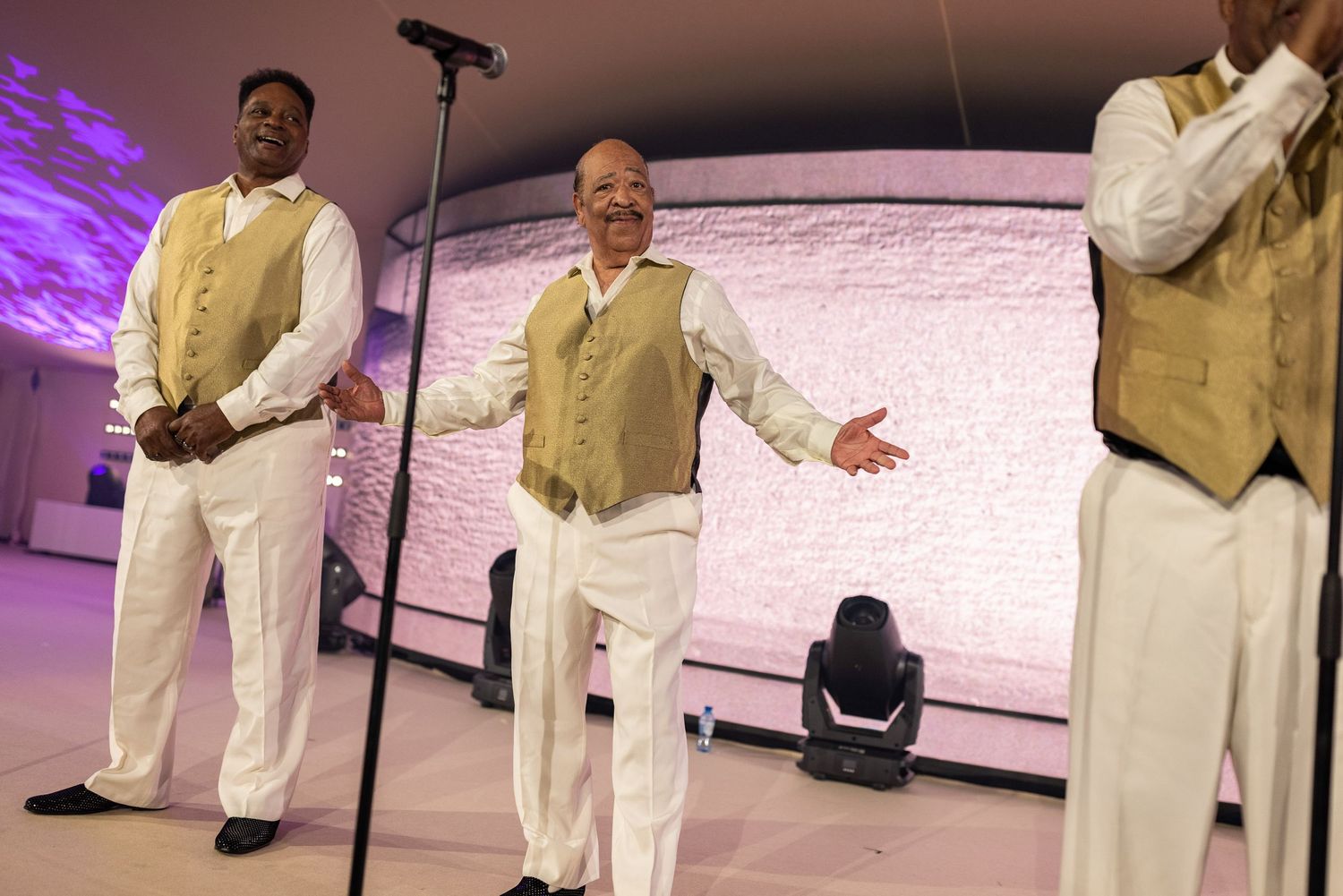 Three performers in matching gold vests and white pants sing on stage with purple lighting and a white backdrop.