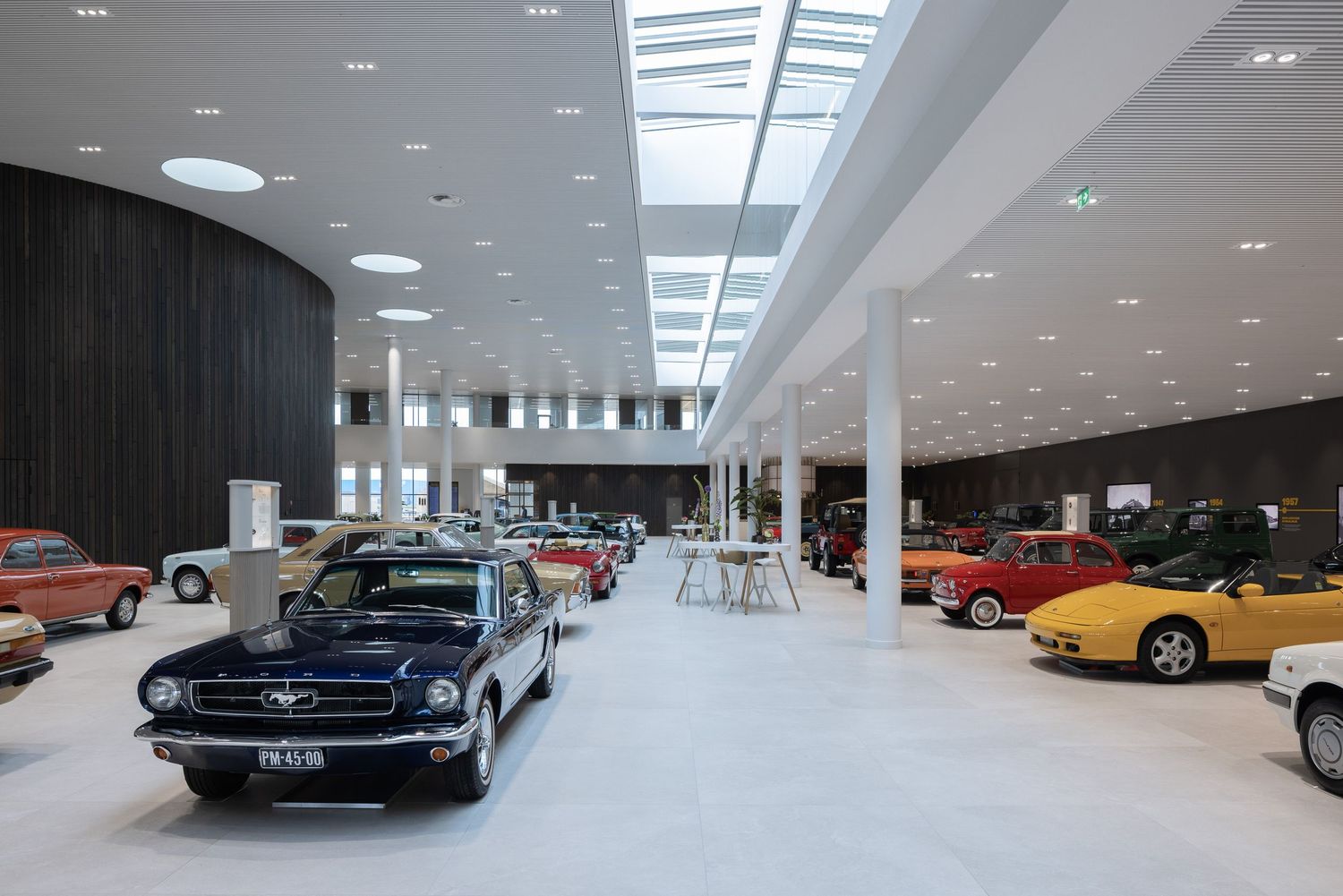 Modern car showroom interior with classic and vintage automobiles displayed under bright skylights and white columns.