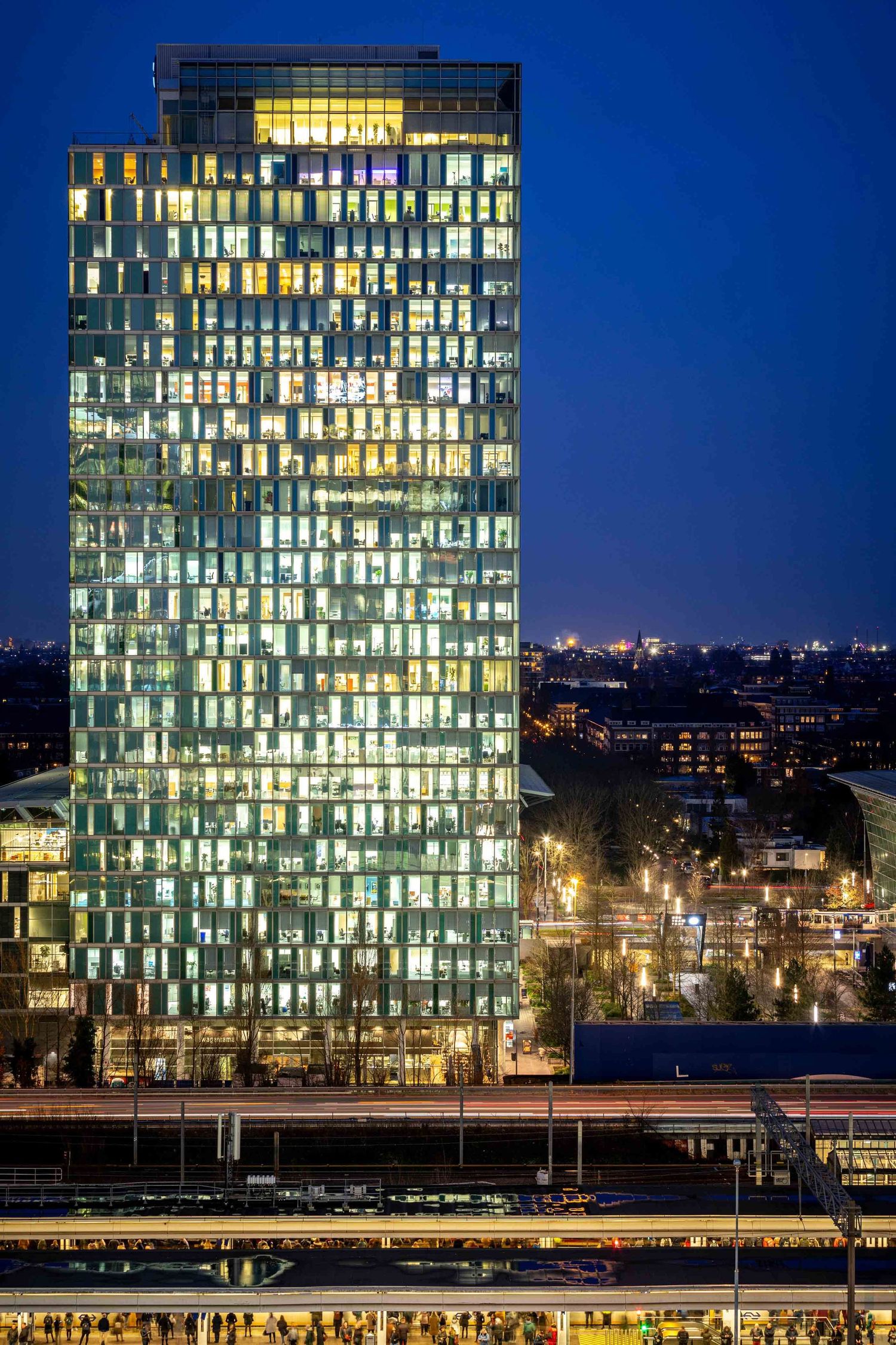 Modern glass office building illuminated at night with glowing windows reflecting against dark blue sky.