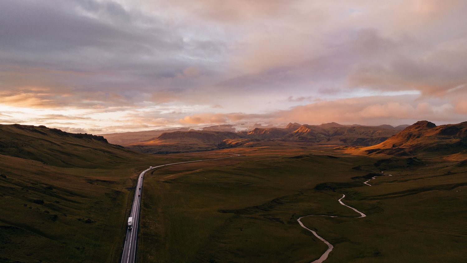 A panoramic view of rolling hills and mountains under dramatic sunset clouds with a winding road and waterfall.