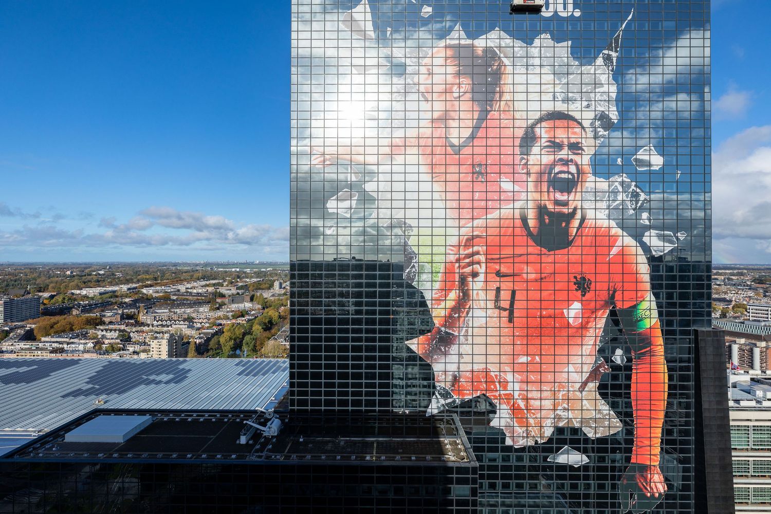 Large mural of a soccer player in orange jersey on a glass skyscraper facade under blue sky.