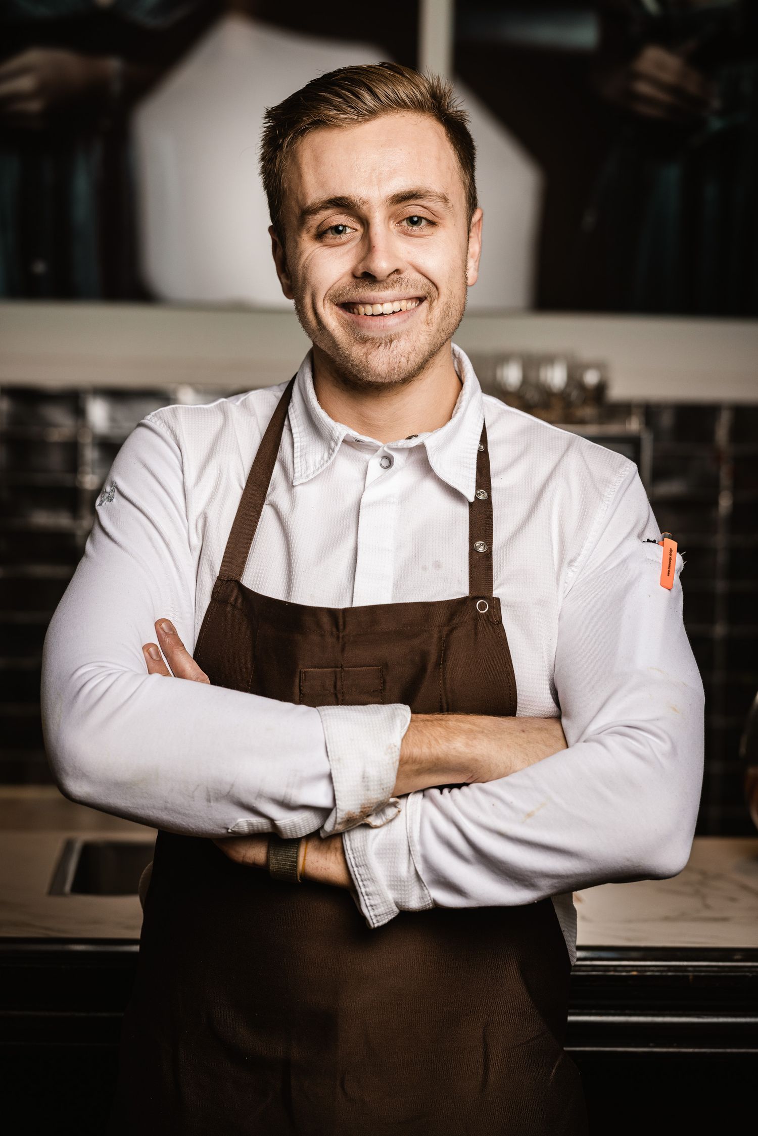 Professional chef in white shirt and brown apron smiling confidently in restaurant kitchen setting.