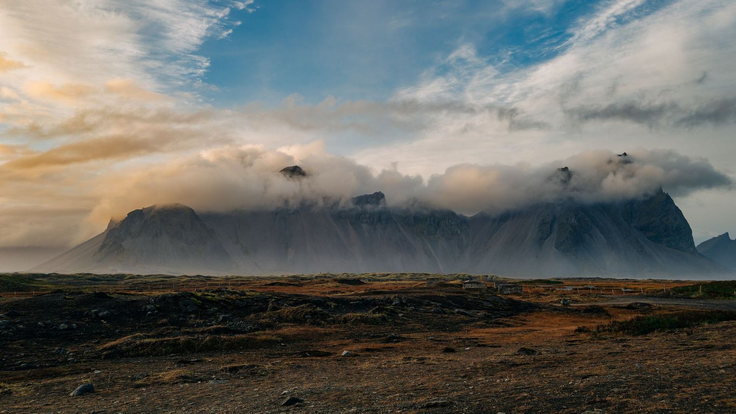 A panoramic view of dramatic mountain landscapes under cloudy skies during sunset with rugged terrain in the foreground.