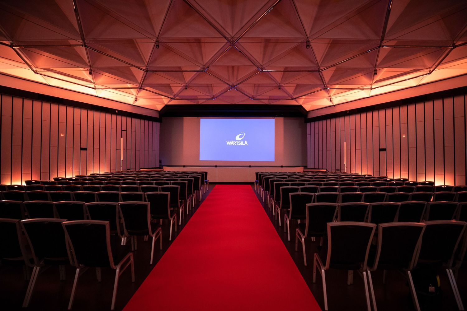 Modern theater auditorium with red seats and dramatic lighting showcasing geometric ceiling design and stage area.
