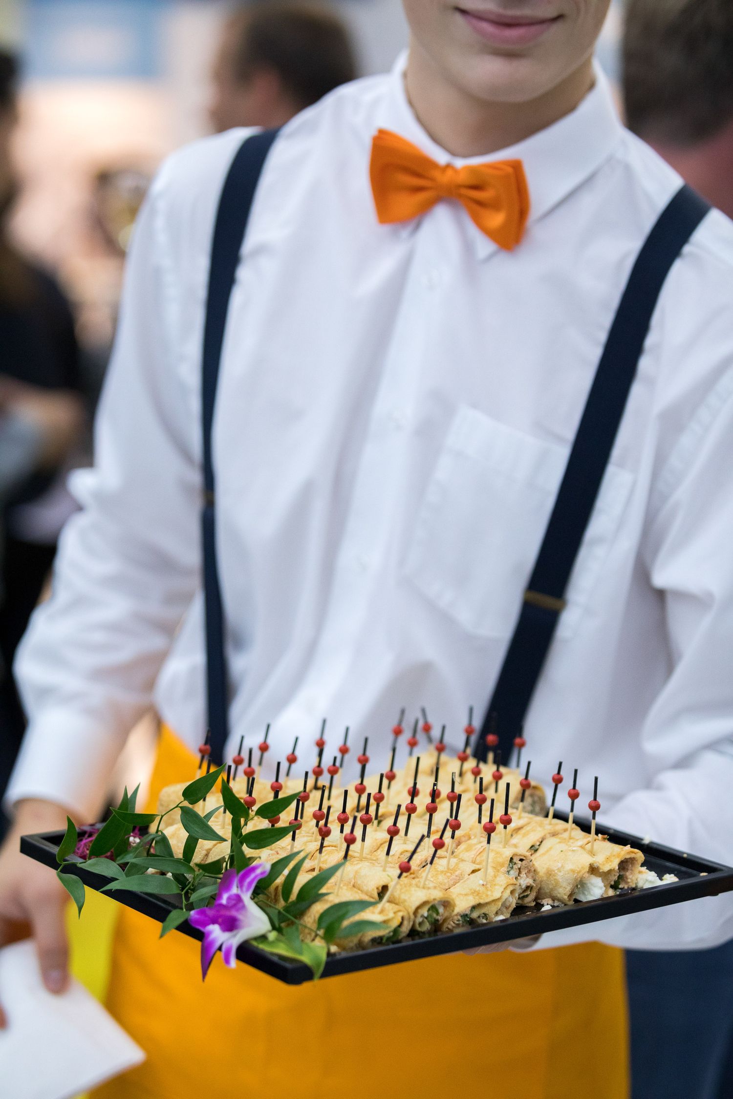 A server wearing suspenders and orange bowtie carries a tray of appetizers decorated with colorful garnish and toothpicks.