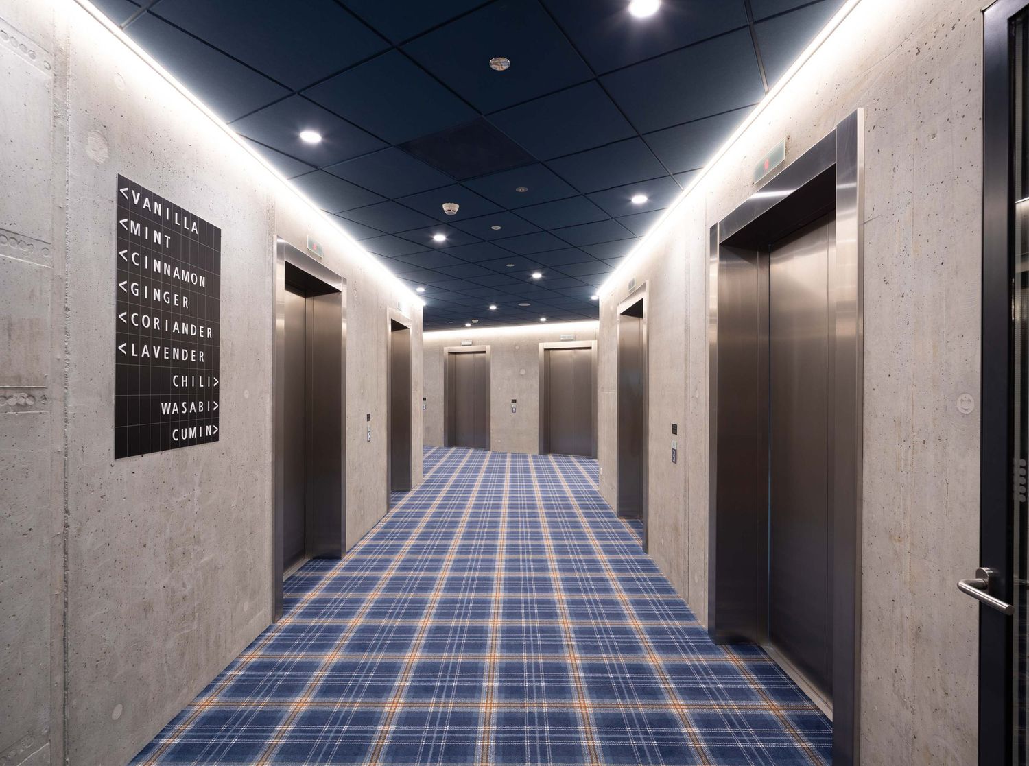 Modern hallway with concrete walls and blue geometric patterned carpet leading to a vanishing point.
