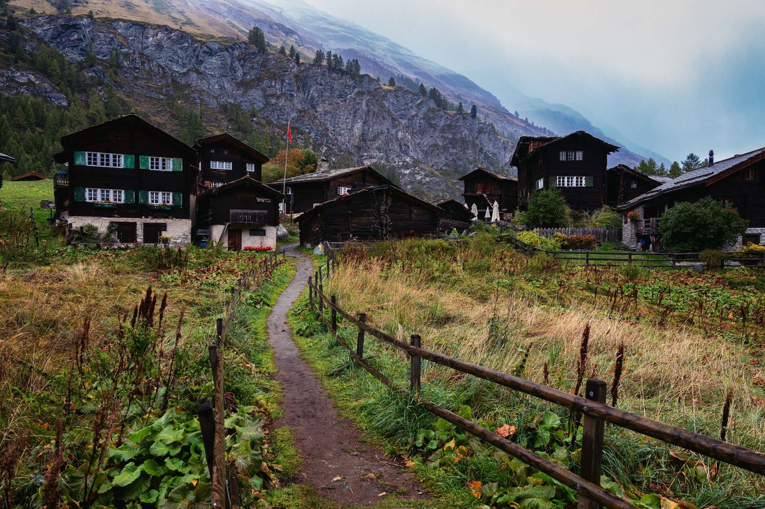 Mountain village path lined with wooden fences and traditional alpine houses against dramatic peaks.