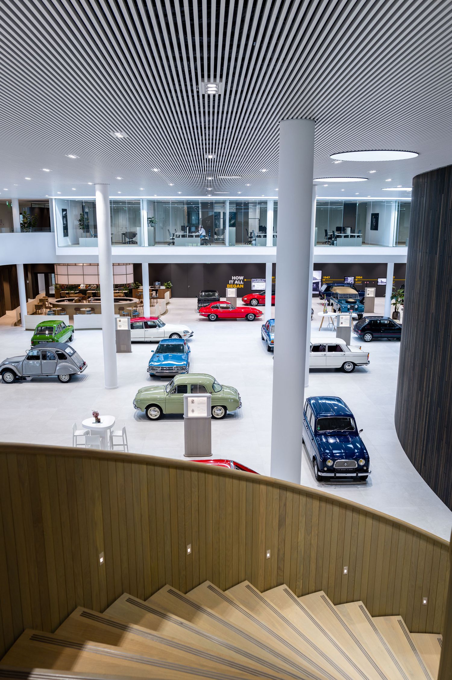 Vintage cars on display in a modern museum showroom with white columns, wood paneling, and descending staircase.
