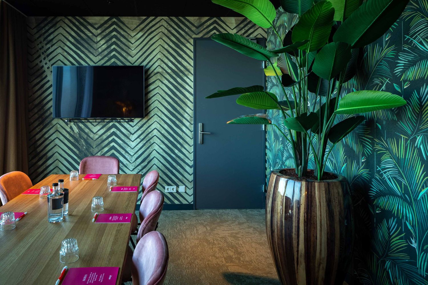 Tropical plants and palm fronds create a lush green atmosphere against a geometric patterned wall in an interior space.