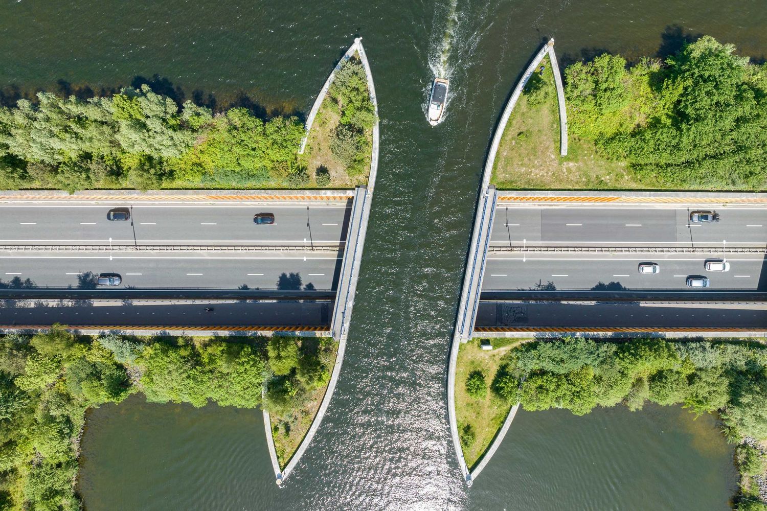 Aerial view of a symmetrical highway bridge split by a narrow waterway, flanked by lush green trees.