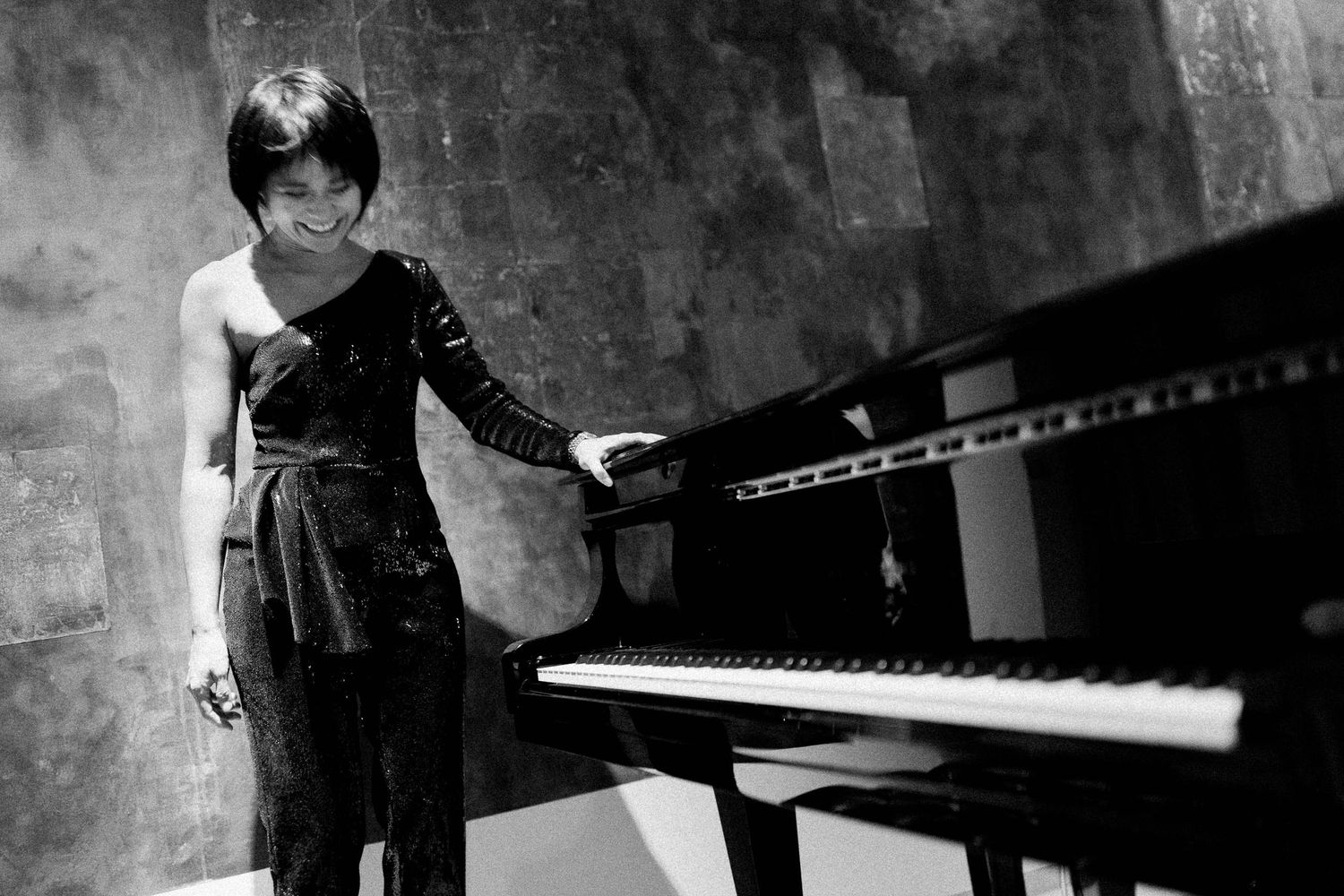 Black and white artistic photograph of a pianist in an elegant dress standing beside a grand piano in moody lighting.