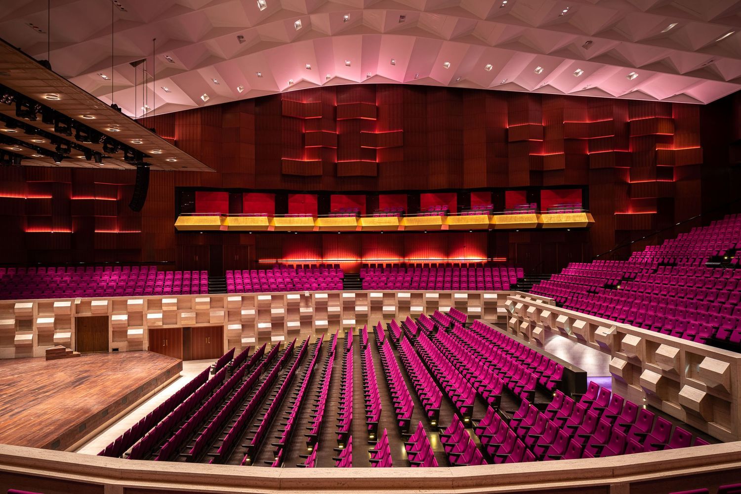 Panoramic view of empty theater auditorium seats arranged in curved rows with magenta lighting and architectural details.