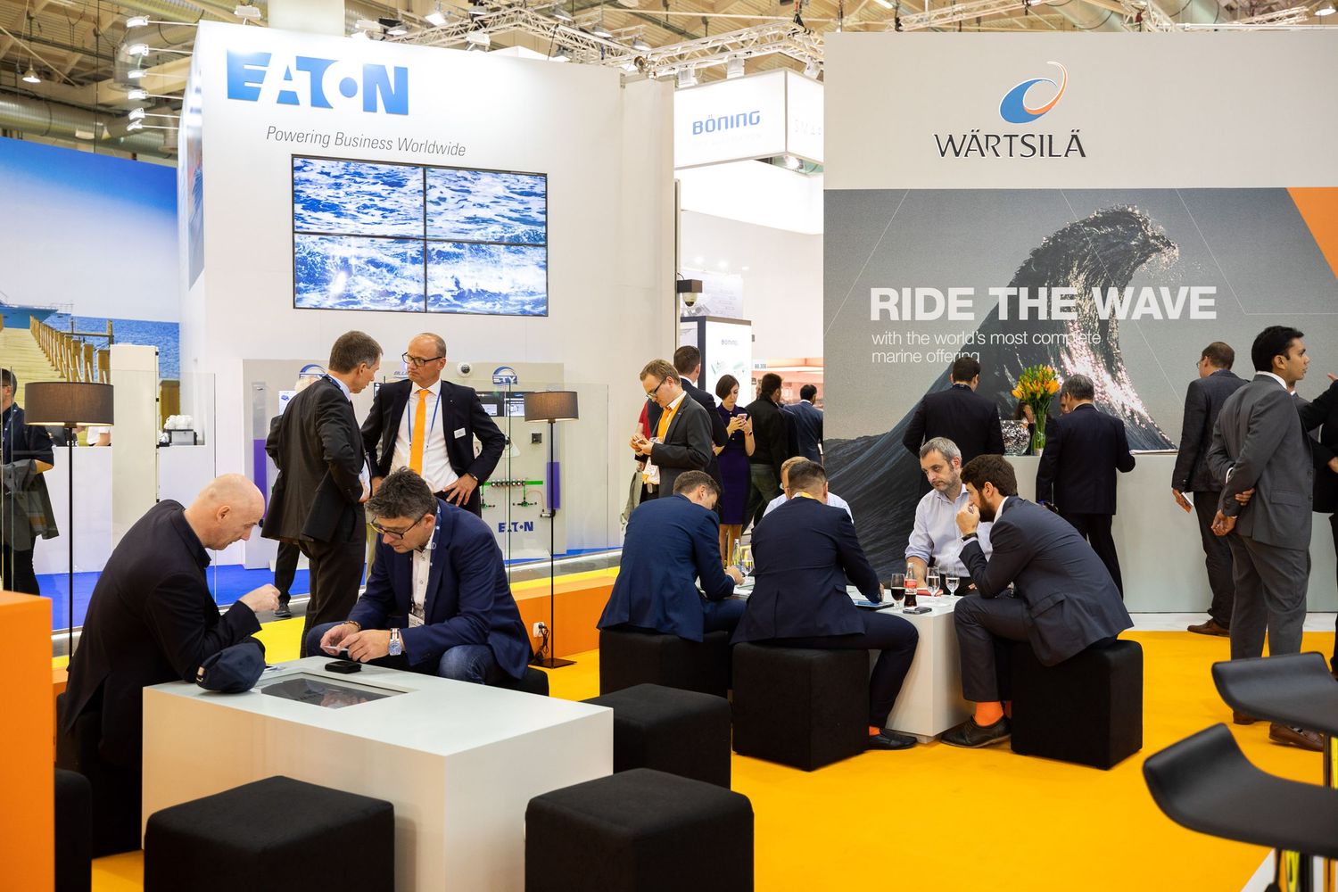 Business exhibition hall with yellow floor displays corporate booths including Eaton and Wartsila company stands.