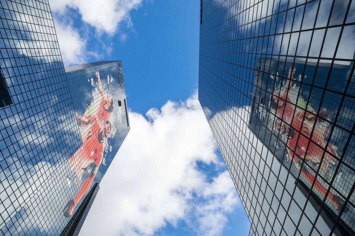 Two glass skyscrapers reflect clouds and a colorful mural under a bright blue sky, viewed from below.