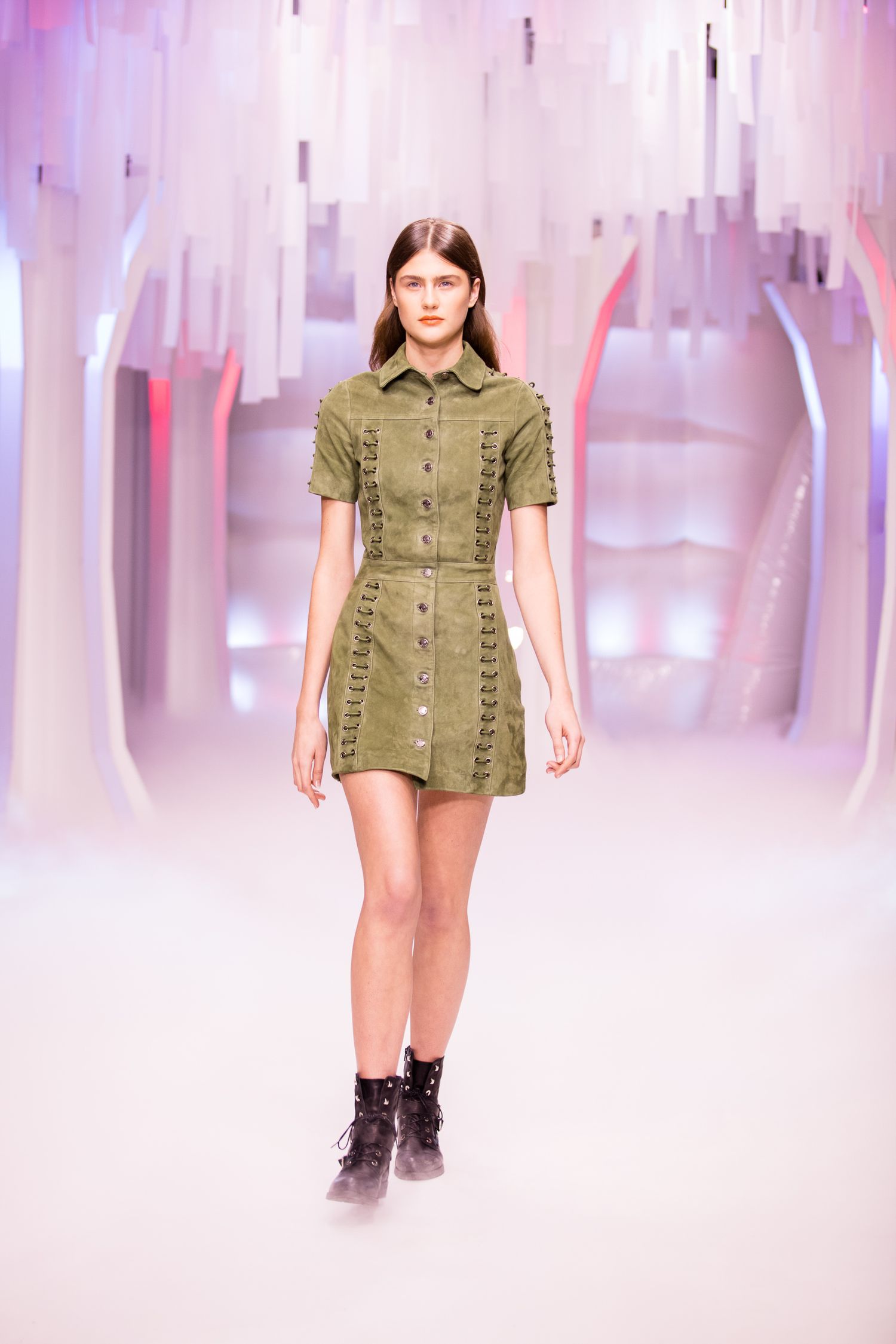 Model walks runway in olive green suede mini dress with lace-up details and black ankle boots at fashion show.