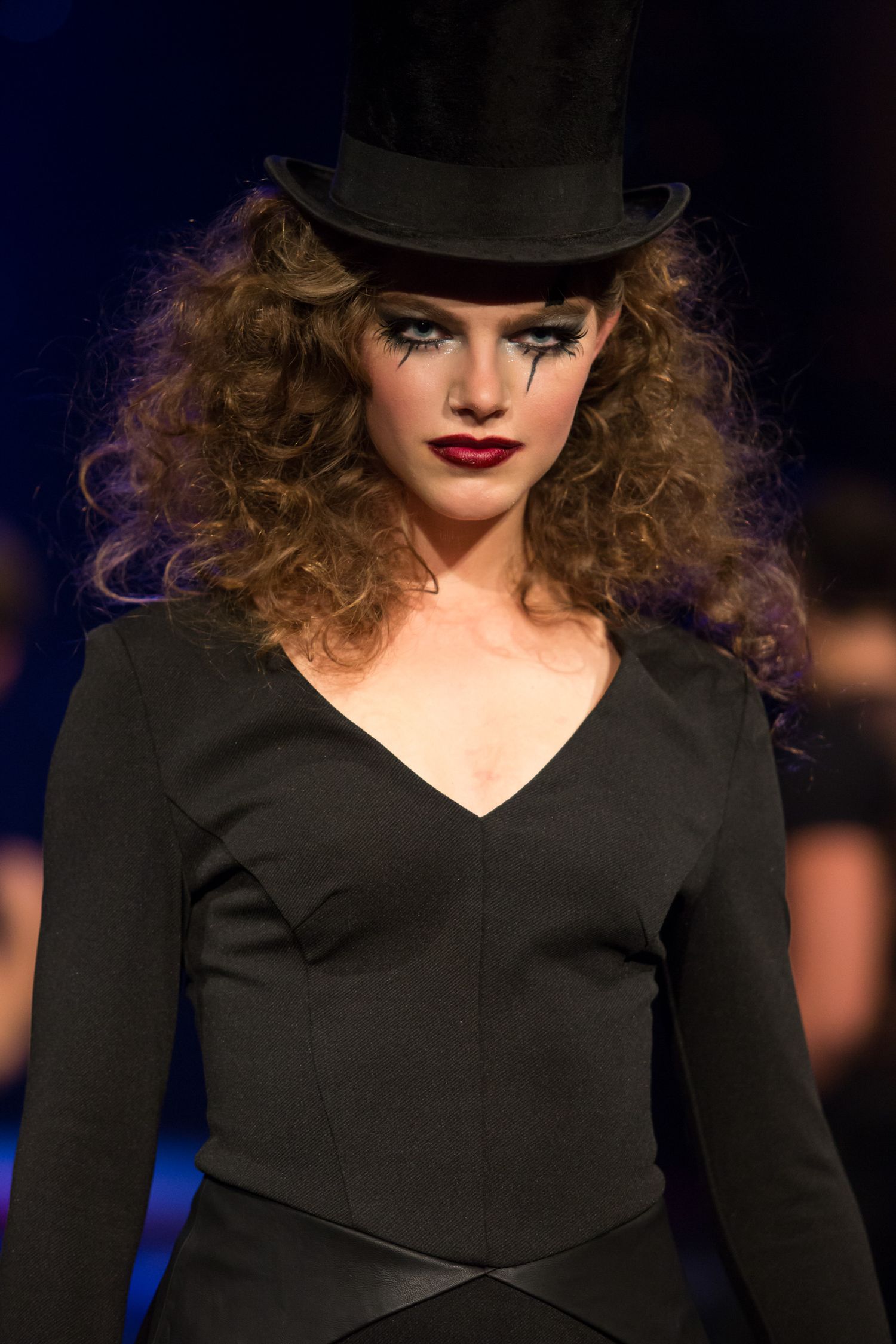 Female model in black top hat and dark outfit with dramatic makeup walks the runway at a fashion show.