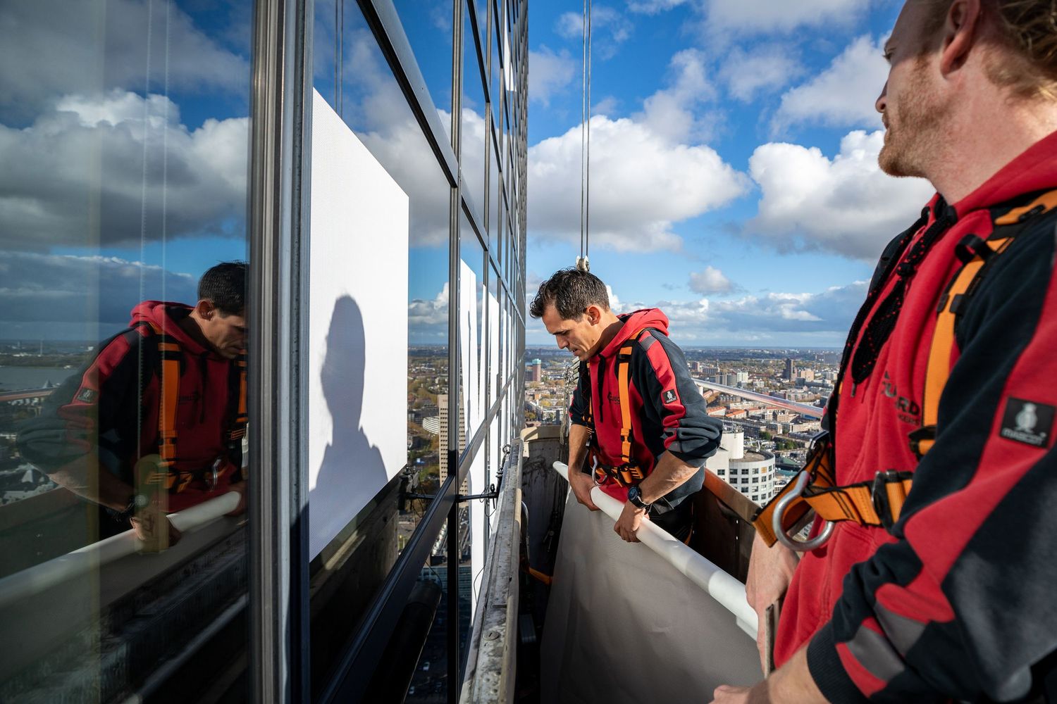Two workers in safety gear inspect high-rise building windows on an elevated platform against a cloudy sky.