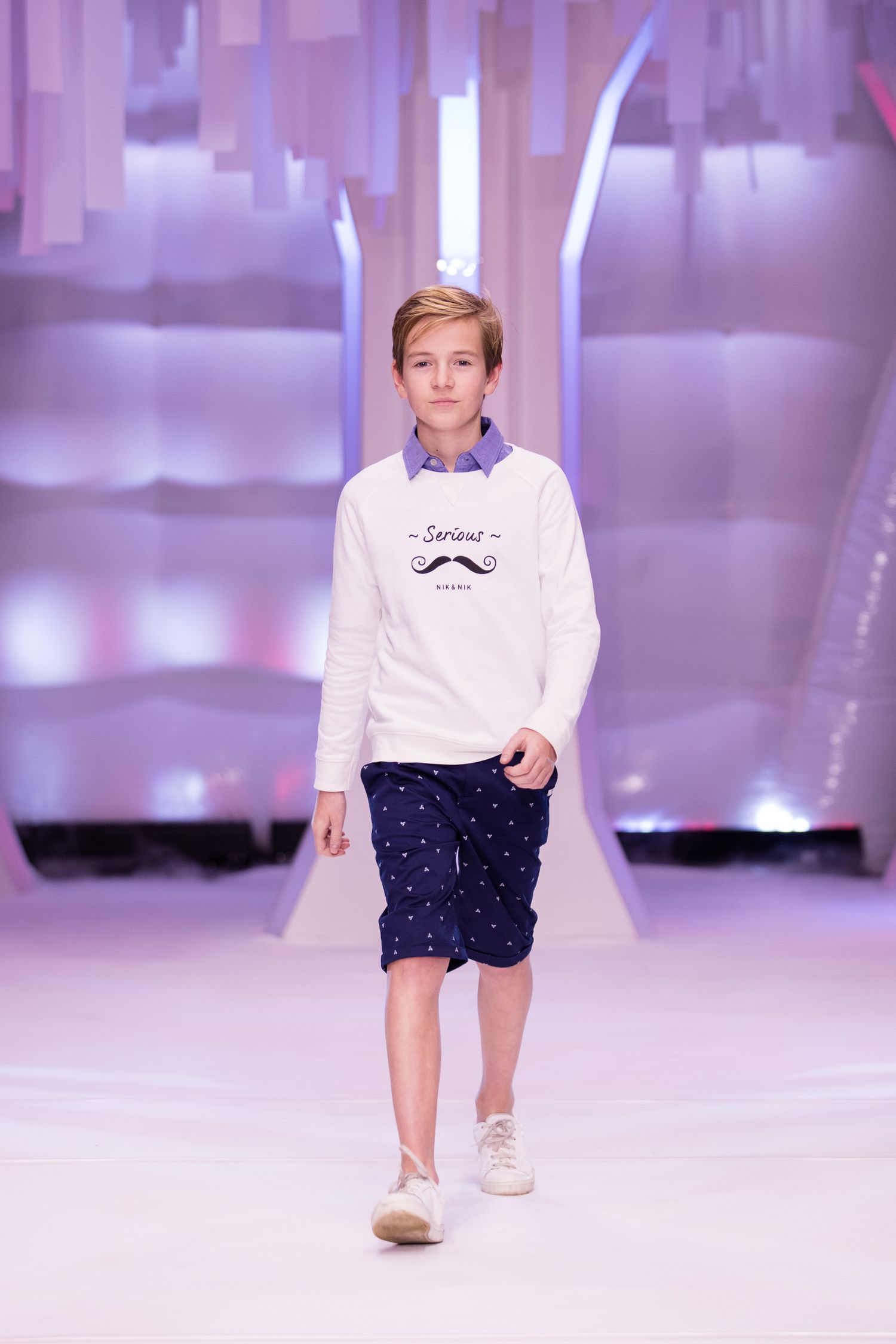 Young boy model walks runway in white mustache sweatshirt, navy dotted shorts, and white sneakers at a fashion show.