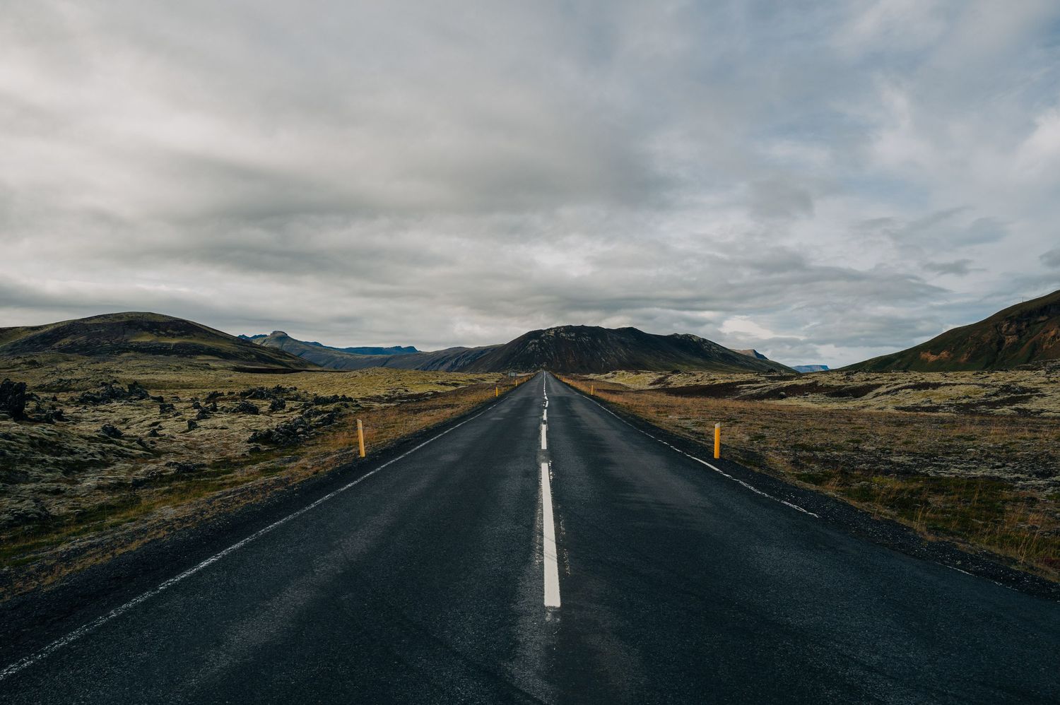 A long empty road stretches through a dramatic Icelandic landscape under moody cloudy skies.
