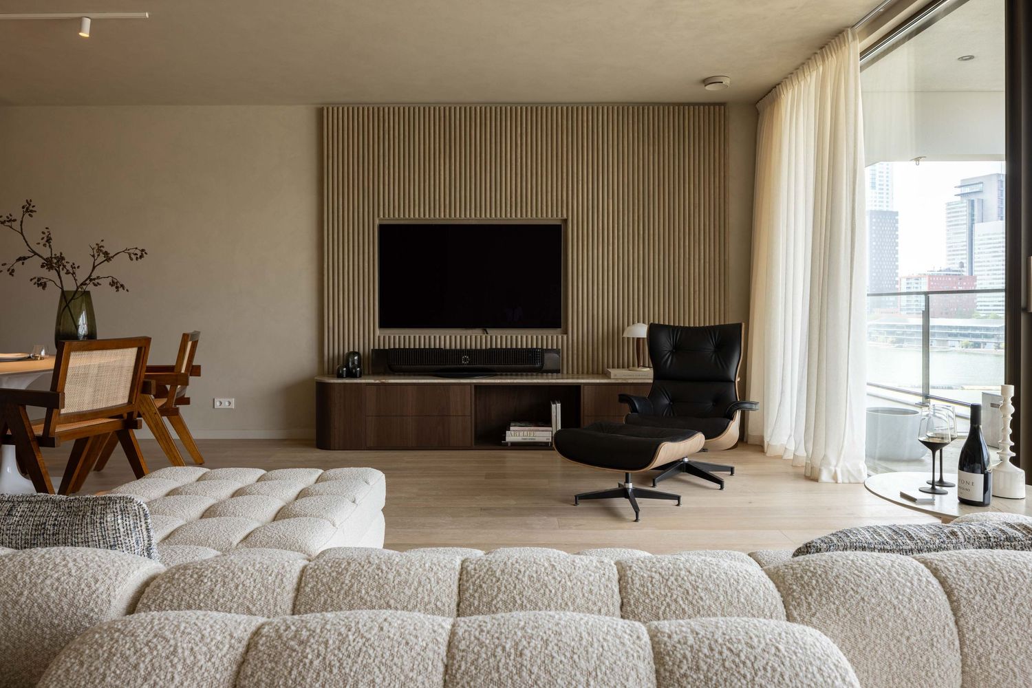 Modern living room with curved beige sectional sofa, wood paneling, and floor-to-ceiling windows.