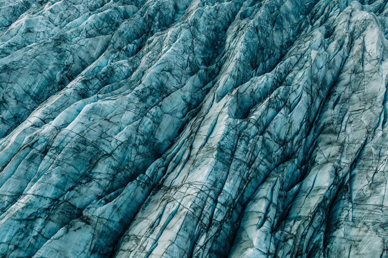 Aerial view of a massive blue glacier with deep crevasses and textured ice formations in a mountainous landscape.