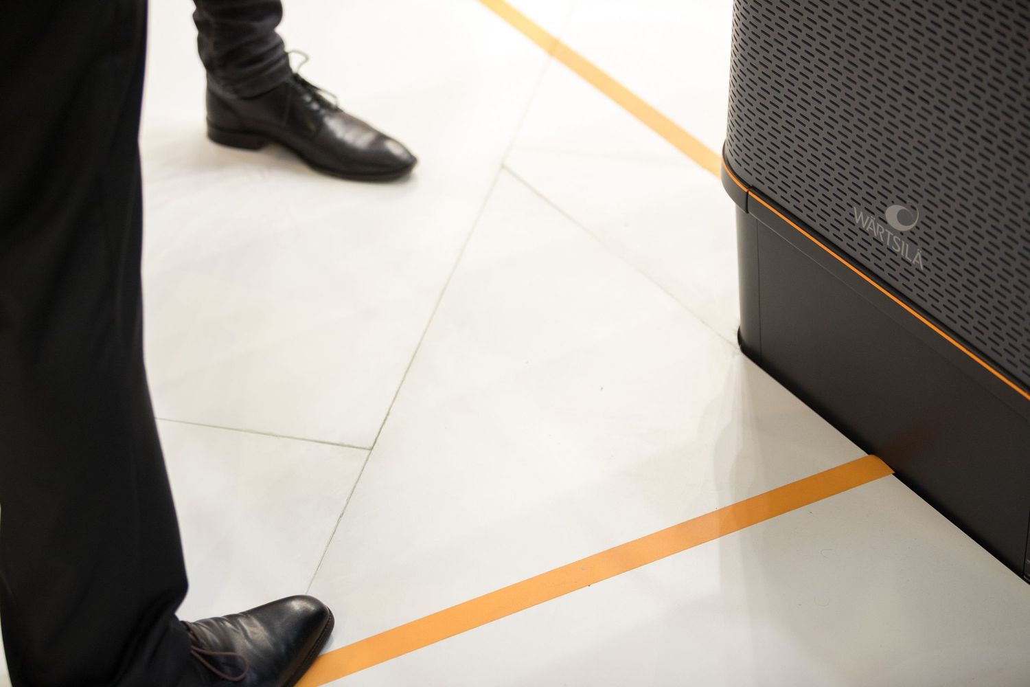 A black dress shoe steps on orange floor tape markings in an office setting.